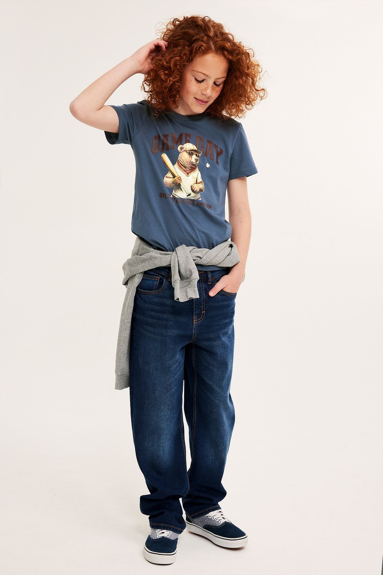 Framifrån: ett barn i blå bomulls-t-shirt med björntryck, mörkblå jeans och blå sneakers.