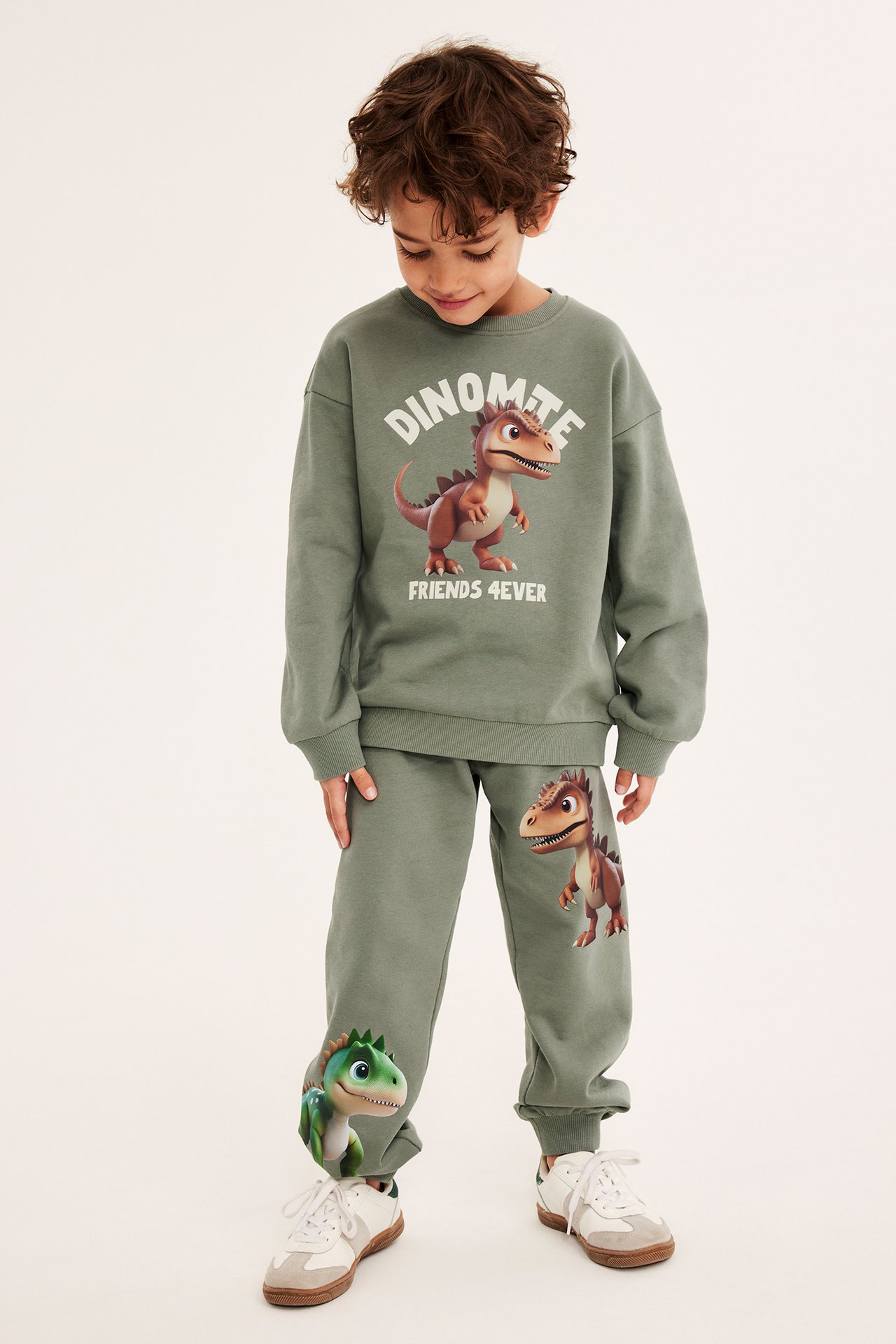 Widok z przodu dziecka w zielonych bawełnianych spodniach dresowych z dinozaurami, pasującej bluzie i białych sneakersach.