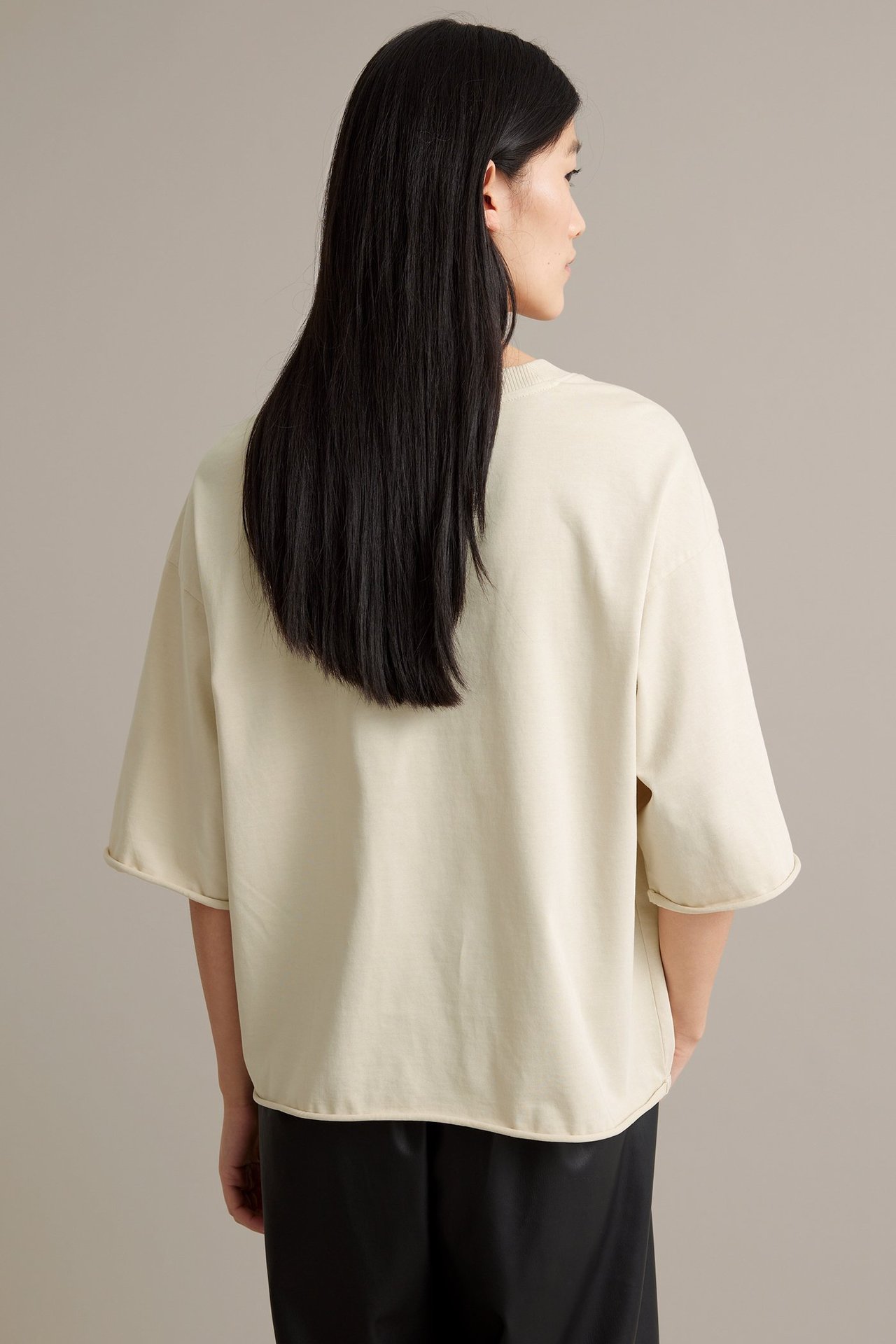 Kvinna i beige T-shirt i grov trikå, bakifrån. Rund hals, råa fållar.