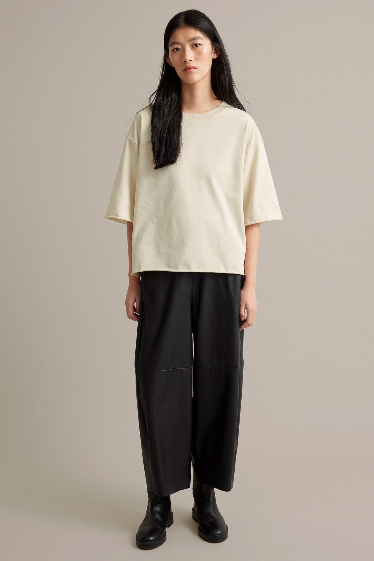 Vaale beige T-paita paksusta trikasta, naisella mustissa housuissa ja saappaissa.