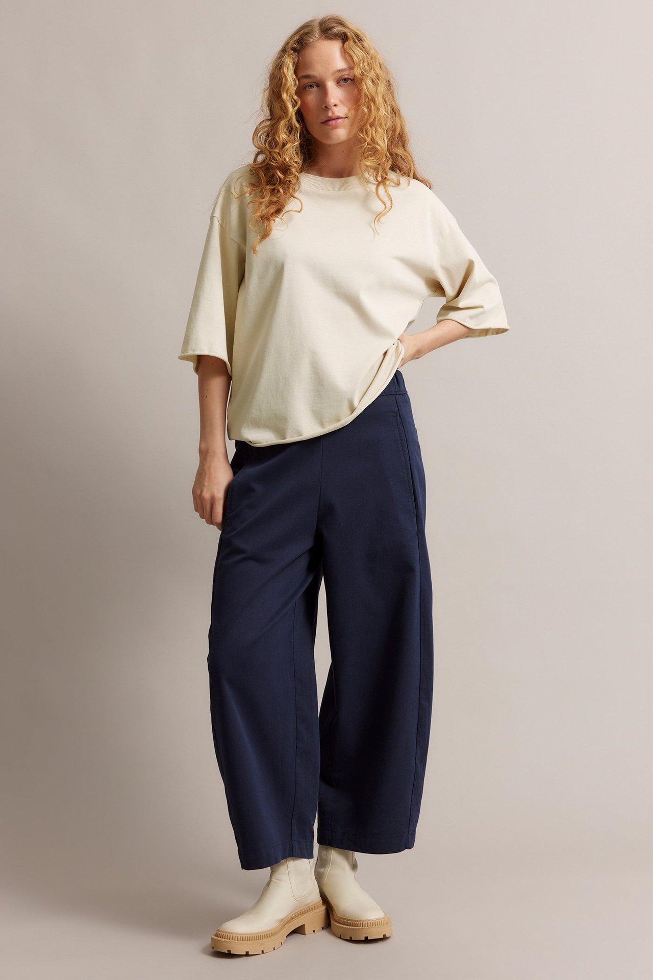 Frontvy av kvinna i ljusbeige bomulls-T-shirt, mörkblå vida byxor och ljusbeige boots.