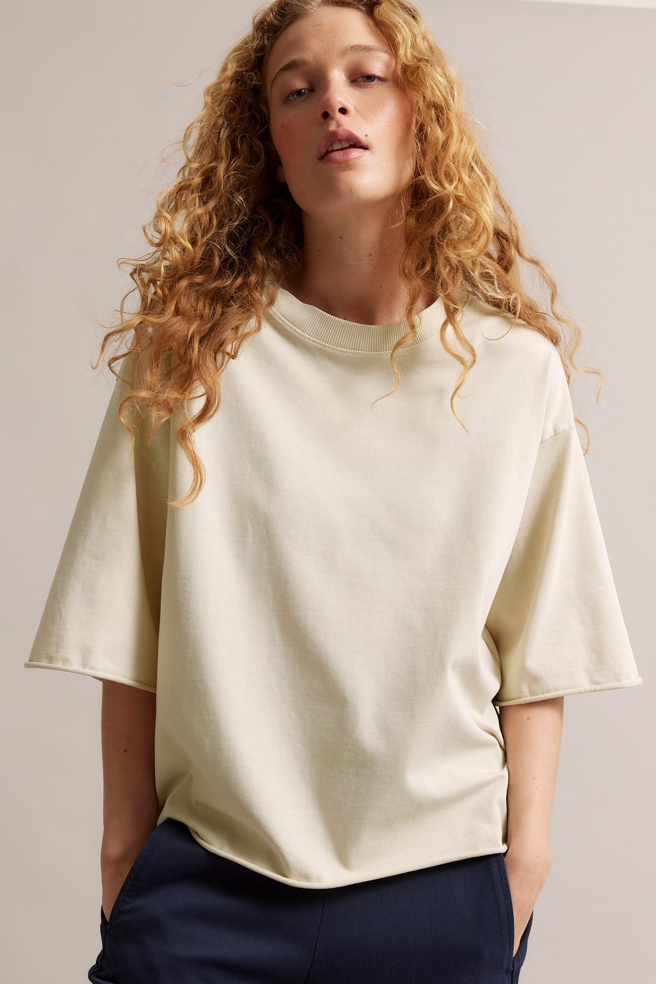 Forfra: Kvinne i lys beige, løs bomull-T-skjorte med korte ermer og råe sømmer, over mørke bukser.