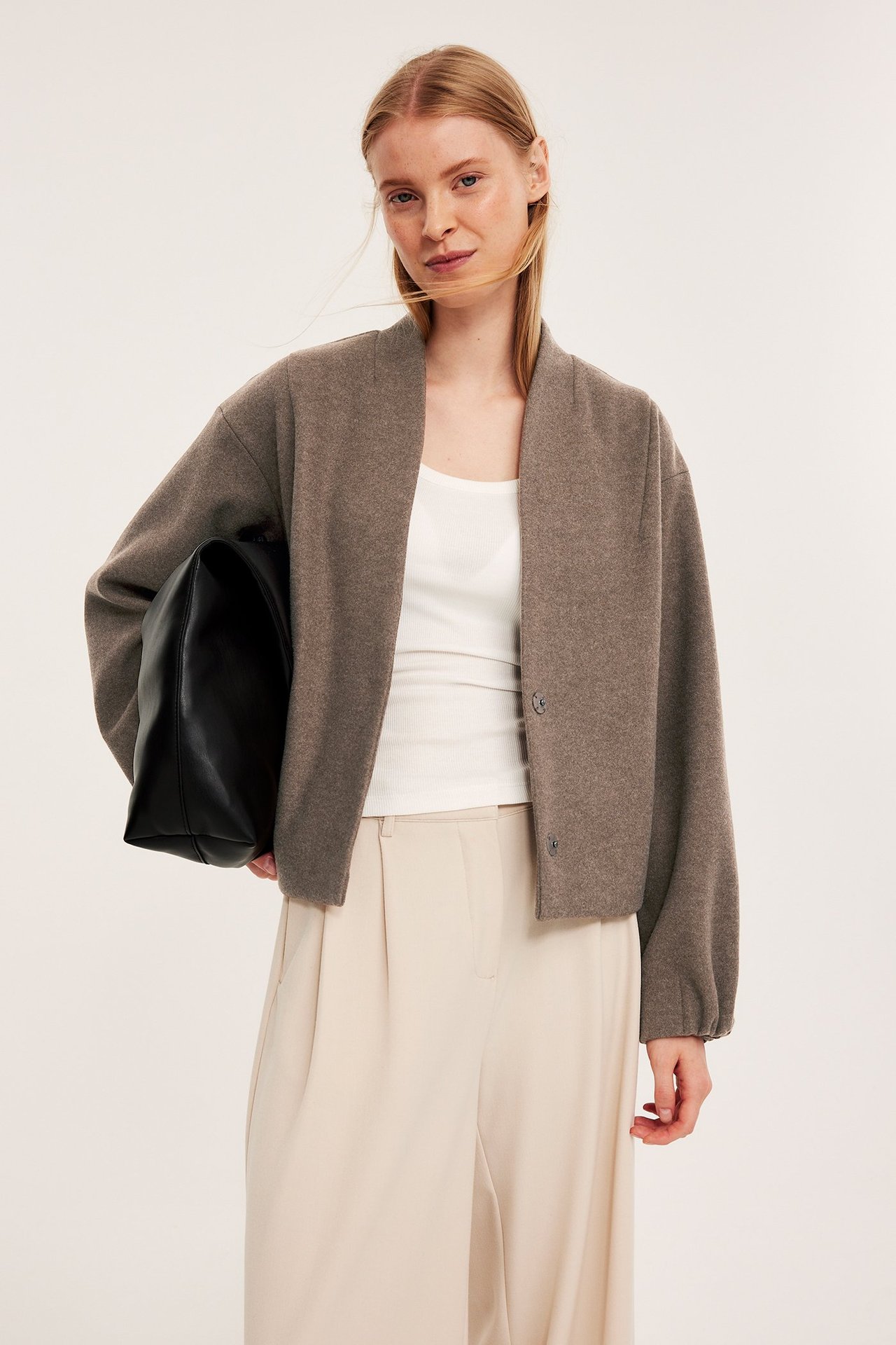 Widok z przodu: Kobieta w brązowej wełnianej kurtce, białym topie, jasnobeżowych spodniach, trzyma czarną torebkę.
