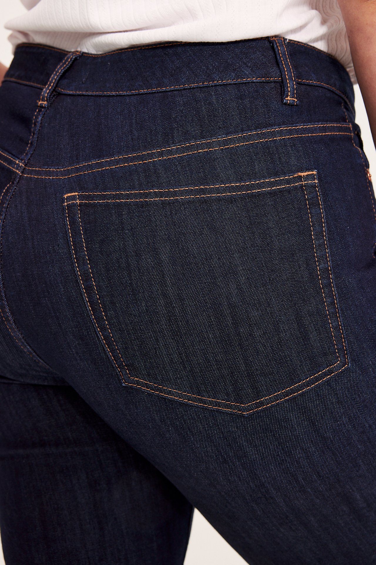 Nærbilde bakfra av en kvinne i mørk high-waist bootcut-jeans med oransje sømmer og hvit topp.