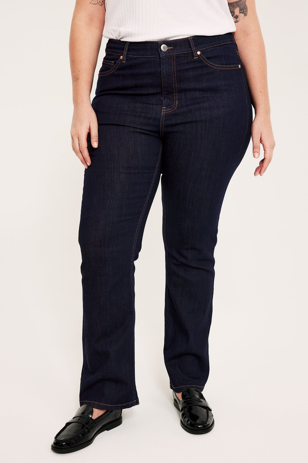 Forfra: kvinne i mørkeblå bootcut-jeans med høyt liv, hvit topp og svarte loafers.