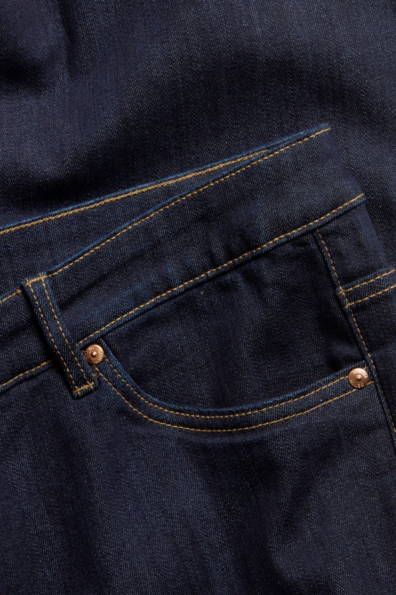 Zbliżenie na ciemnoniebieskie jeansy bootcut z żółtymi szwami i miedzianymi nitami na przedniej kieszeni.