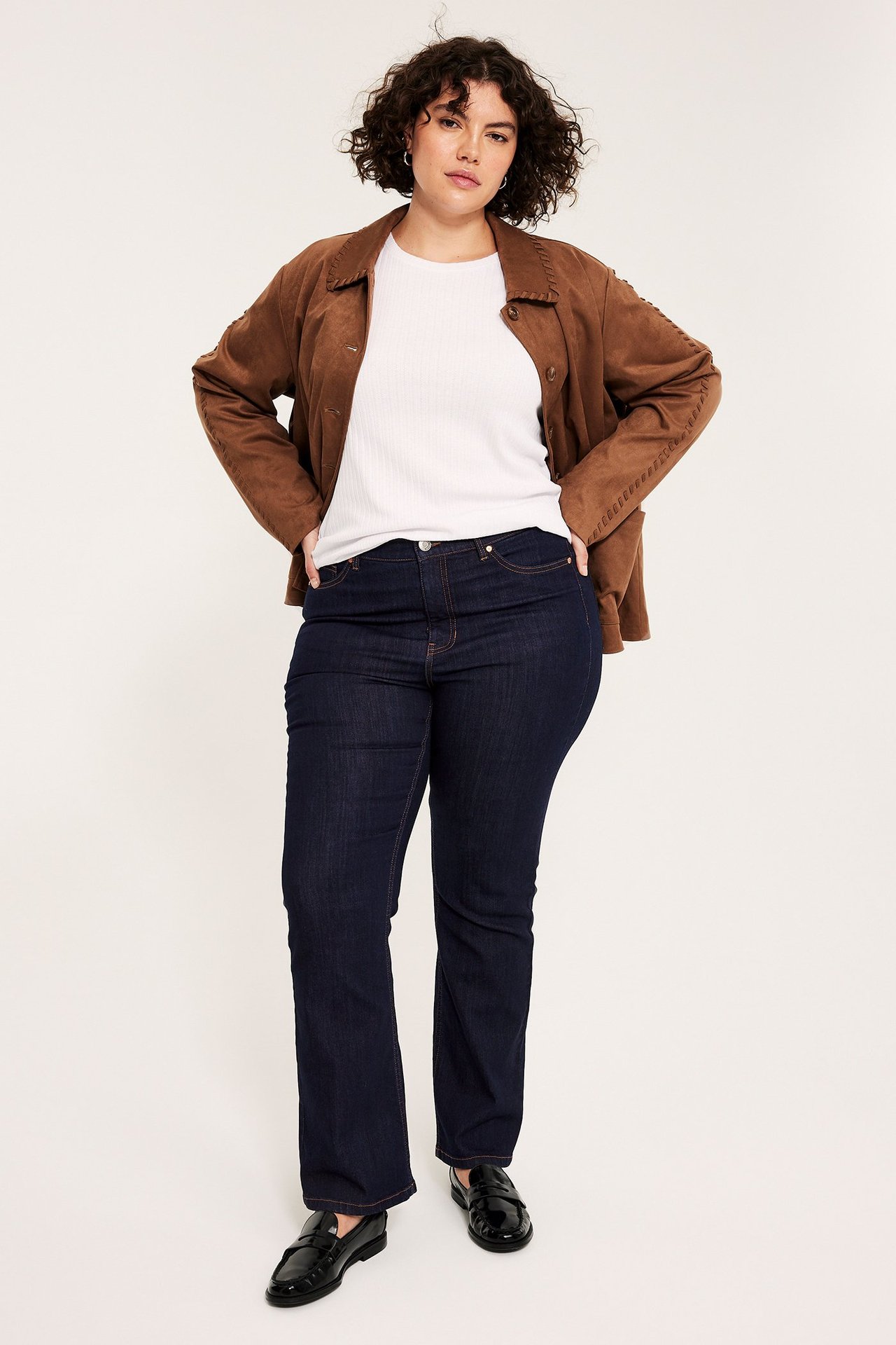 Edestäpäin: nainen yllään tummansiniset korkeavyötäröiset bootcut-farkut, valkoinen ribbipaita ja ruskea takki.