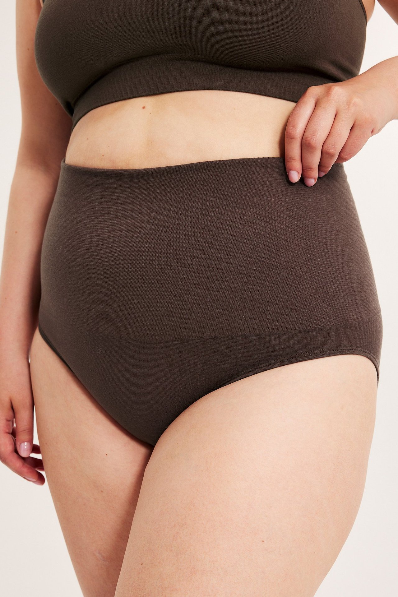 Nainen pukeutuu tummanruskeisiin saumattomiin korkeavyötäröisiin alushousuihin ja vastaavaan braletteen. Käsi vyötäröllä.