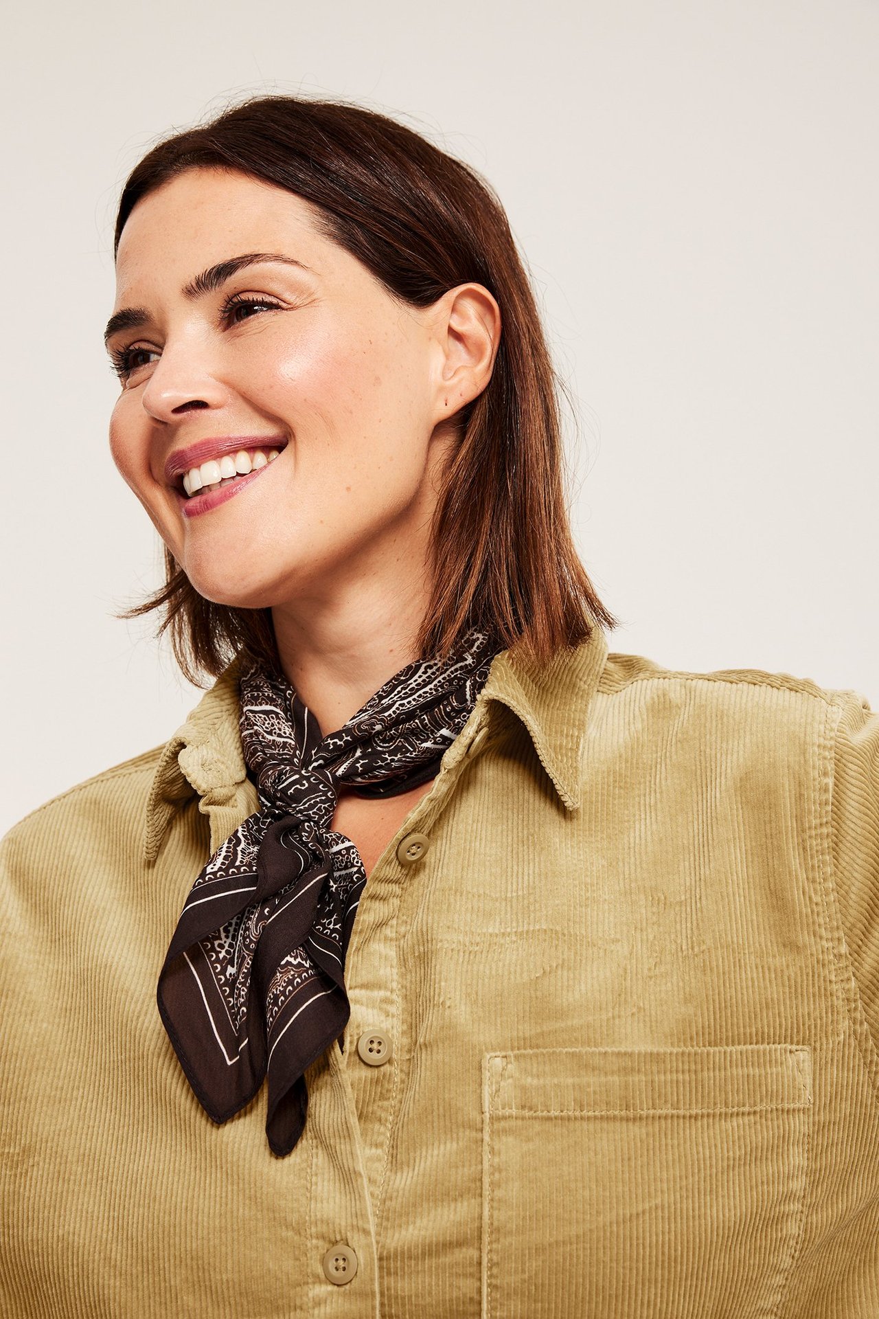 Lähikuva naisesta tummanbeigessä vakosamettipaidassa, musta kuviollinen huivi kaulan ympärillä.