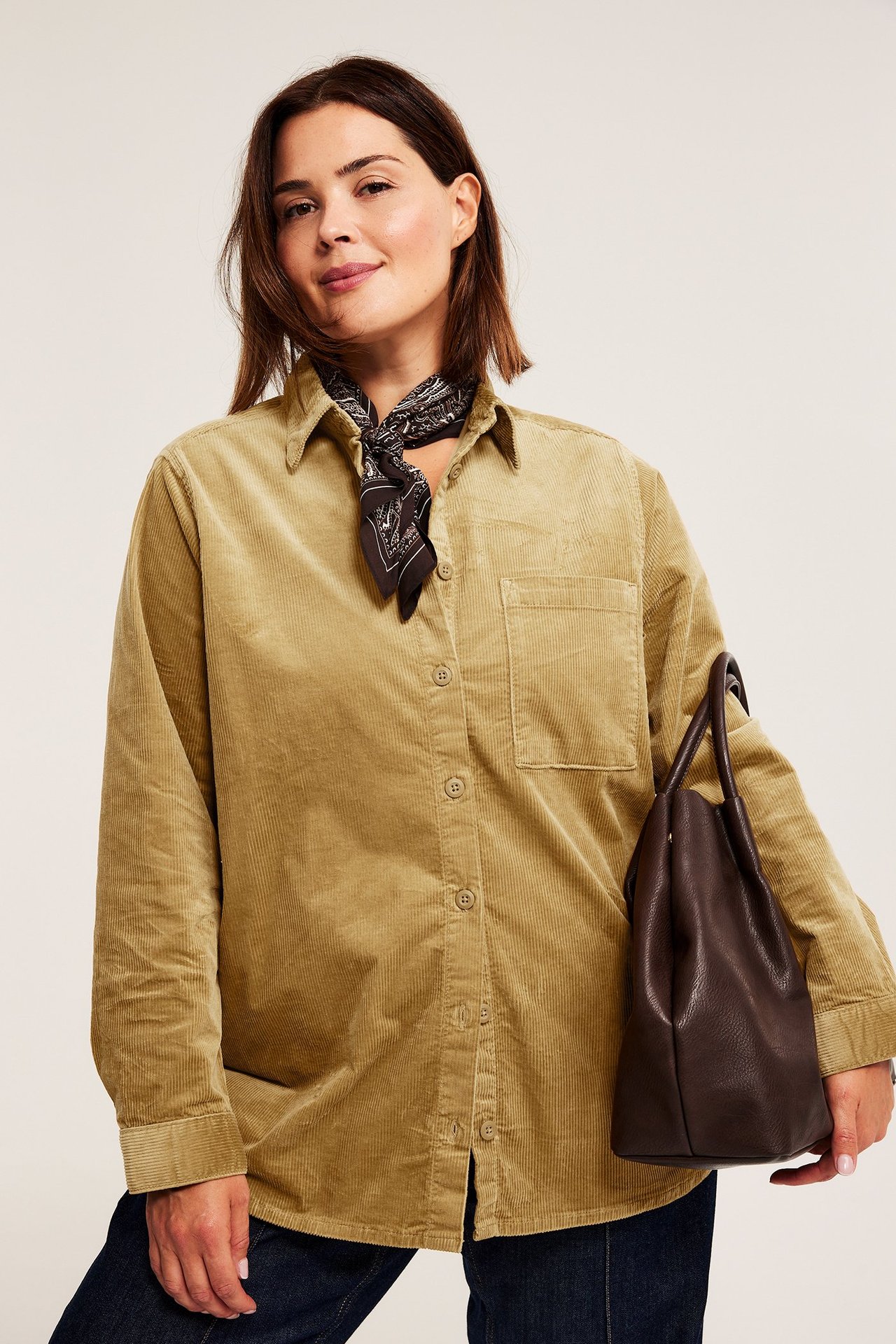 Edestä: Nainen beigessä vakosamettipaidassa, kuviollinen huivi, tummat farkut, pitää ruskeaa käsilaukkua.