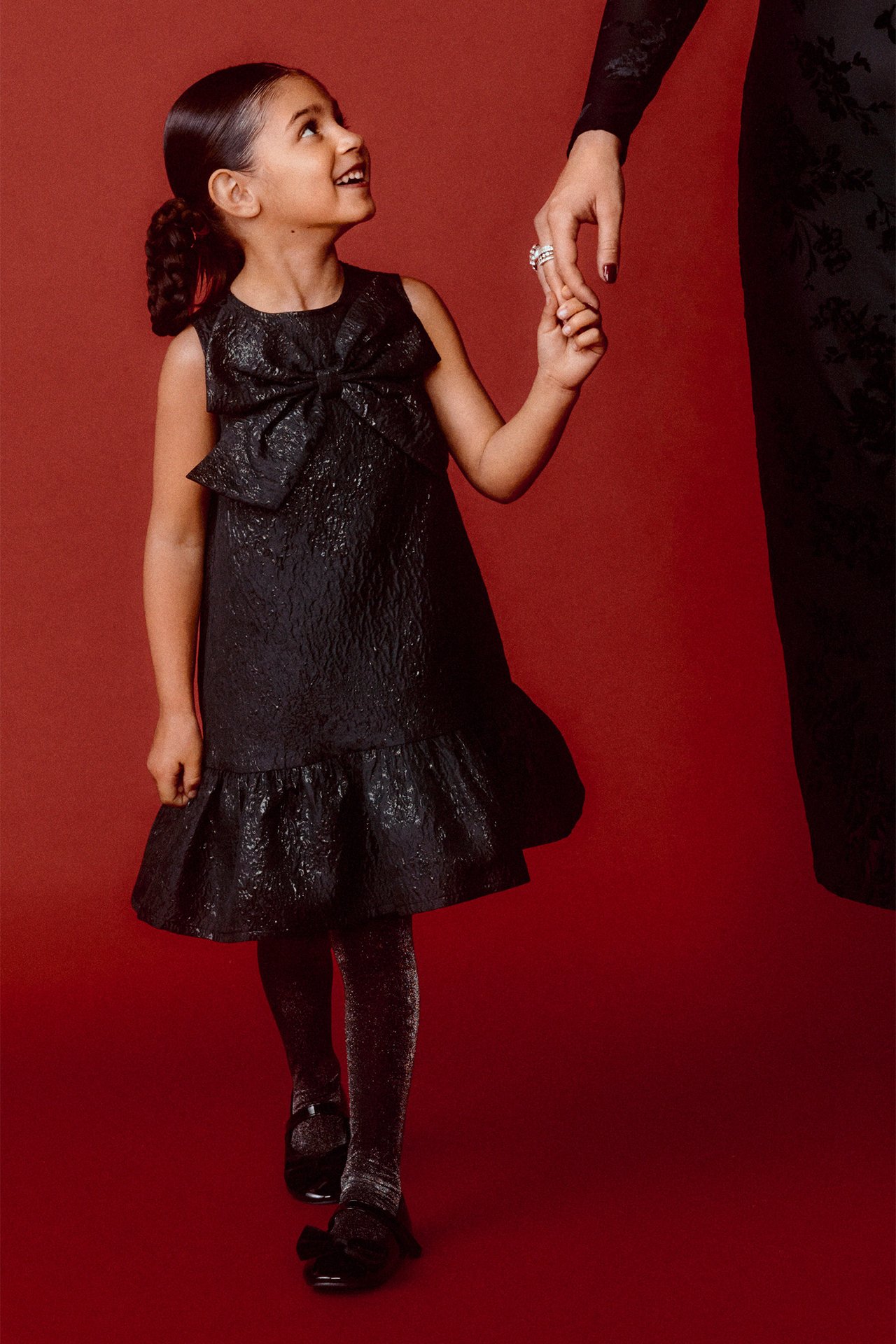 Barn i svart brokadklänning med rosett fram, glittriga strumpbyxor och svarta ballerinaskor, hållande kvinnas hand.