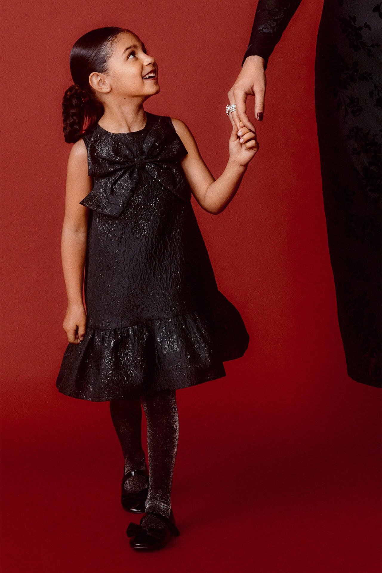 Barn i svart brokadklänning med rosett fram, glittriga strumpbyxor och svarta ballerinaskor, hållande kvinnas hand.