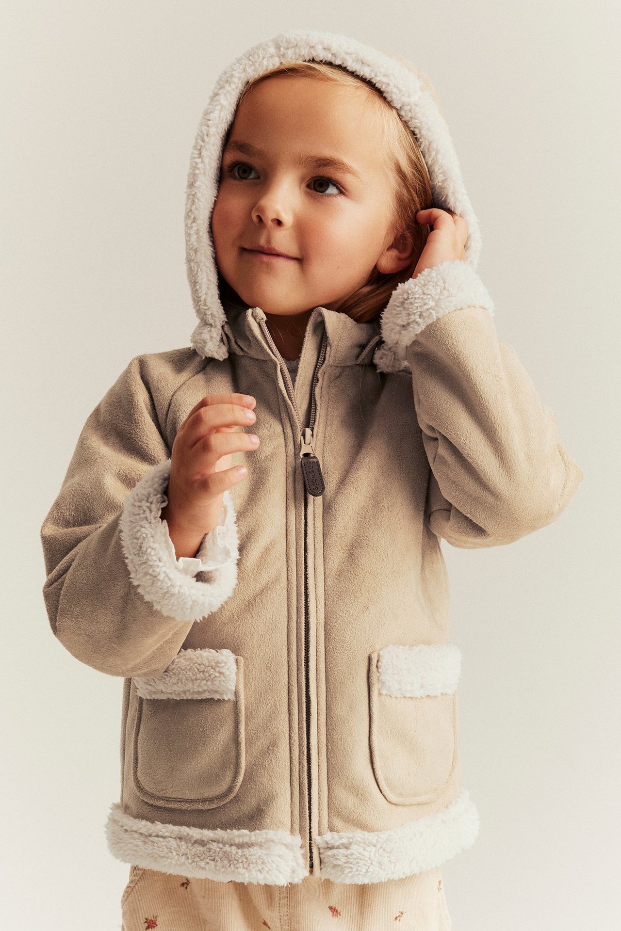 Edestä, lapsi beigessä shearling-tyyppisessä takissa pörrövuorilla ja vaaleissa kukkakuvioisissa housuissa.