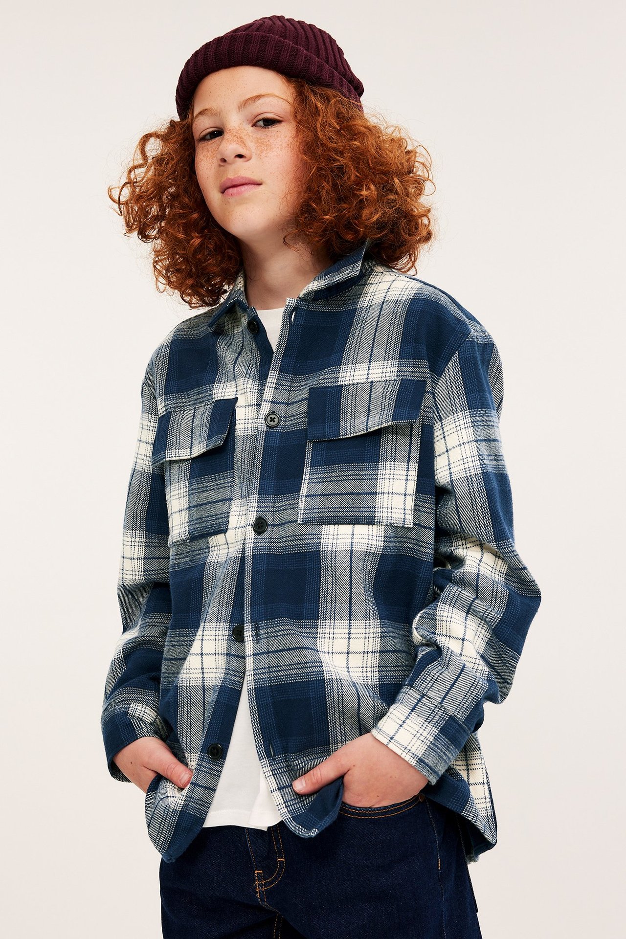 Framifrån: Barn i mörkblårutig flanellöverskjorta över vit t-shirt, mörka jeans och mörkröd mössa.