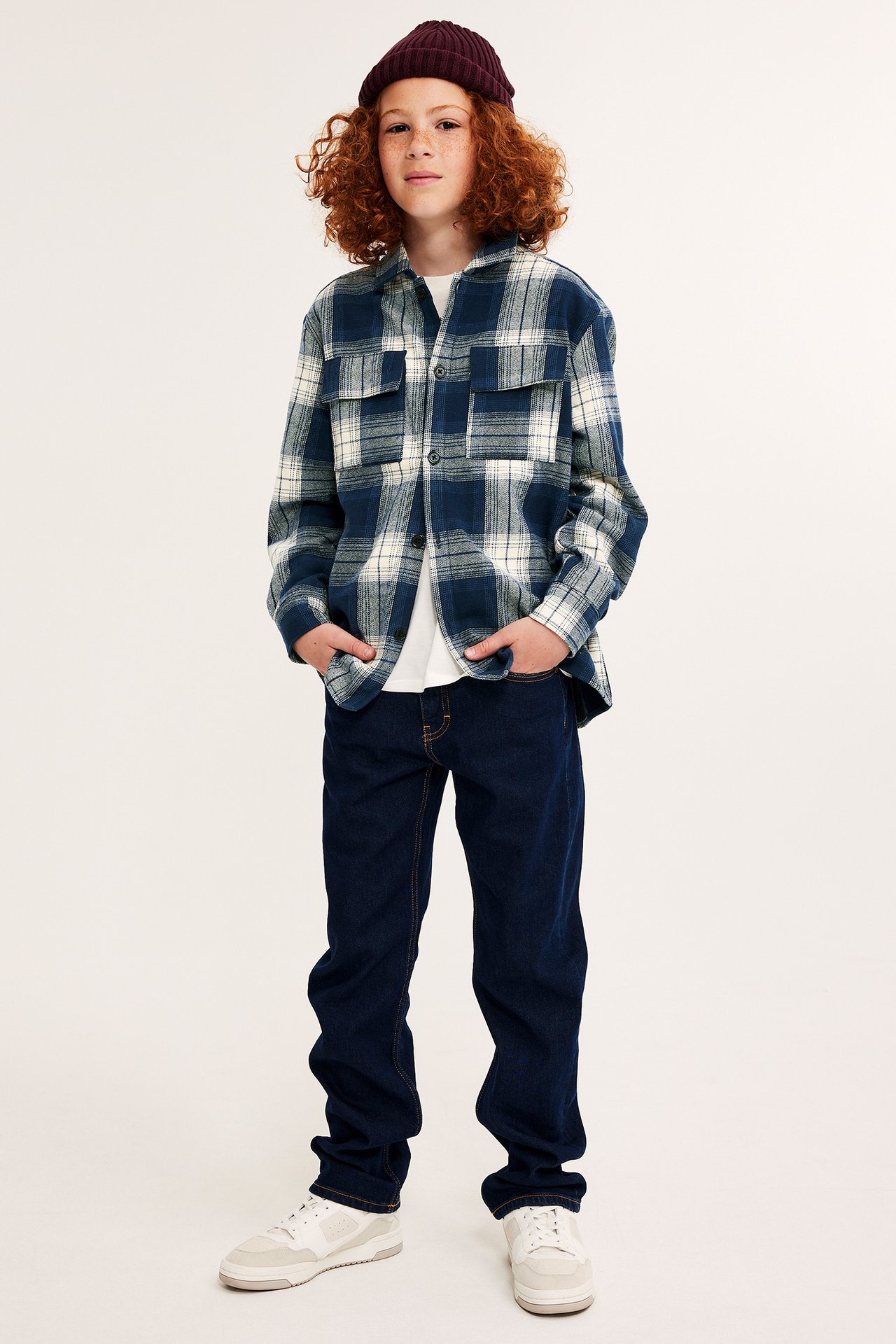Barn i mørkeblå rutete flanellskjorte, hvit t-skjorte, mørkeblå jeans, hvite joggesko og mørkerød lue.