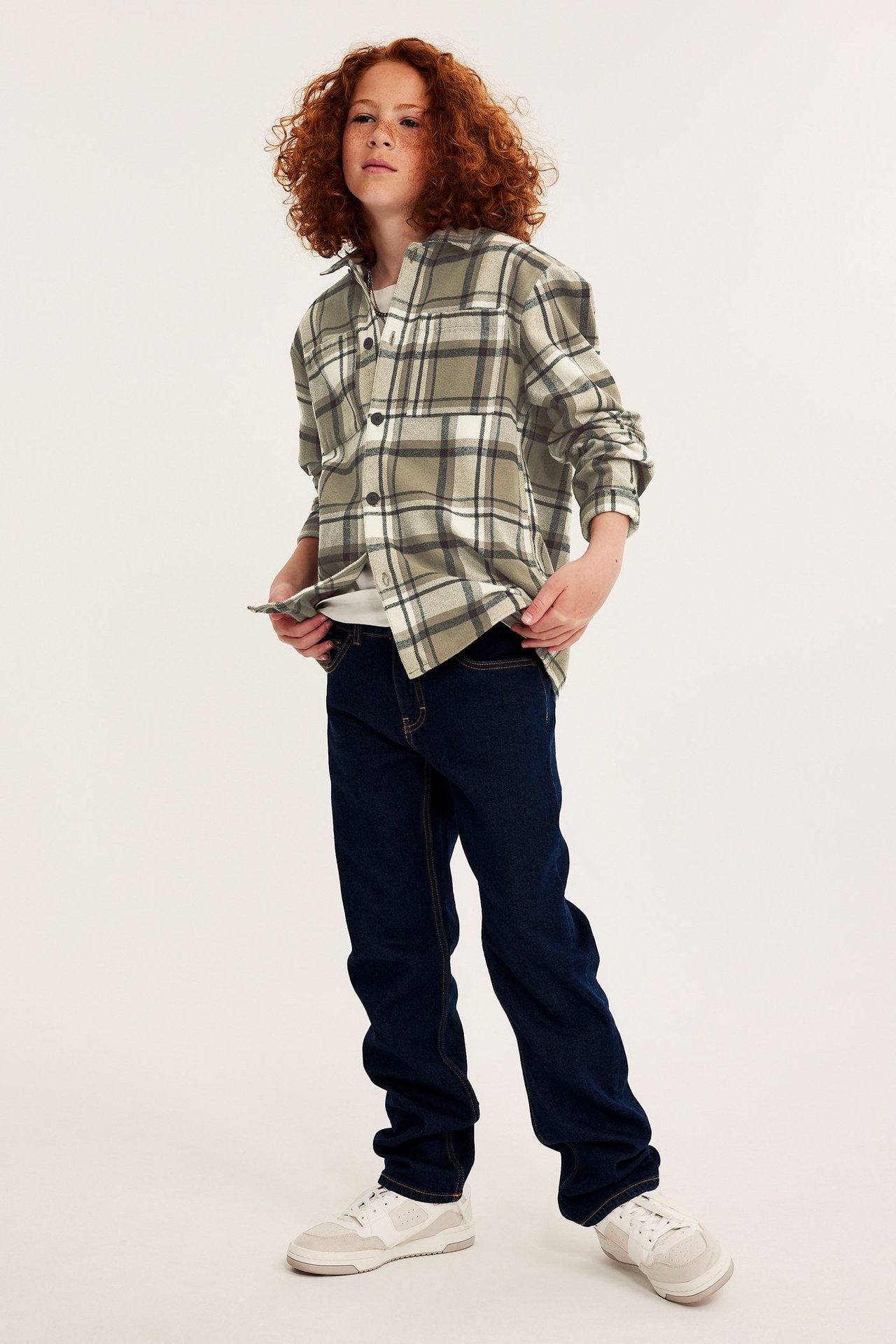 Frontvy av ett barn i mörkbeige rutig flanellöverskjorta, vit t-shirt, mörkblå jeans, vita sneakers.
