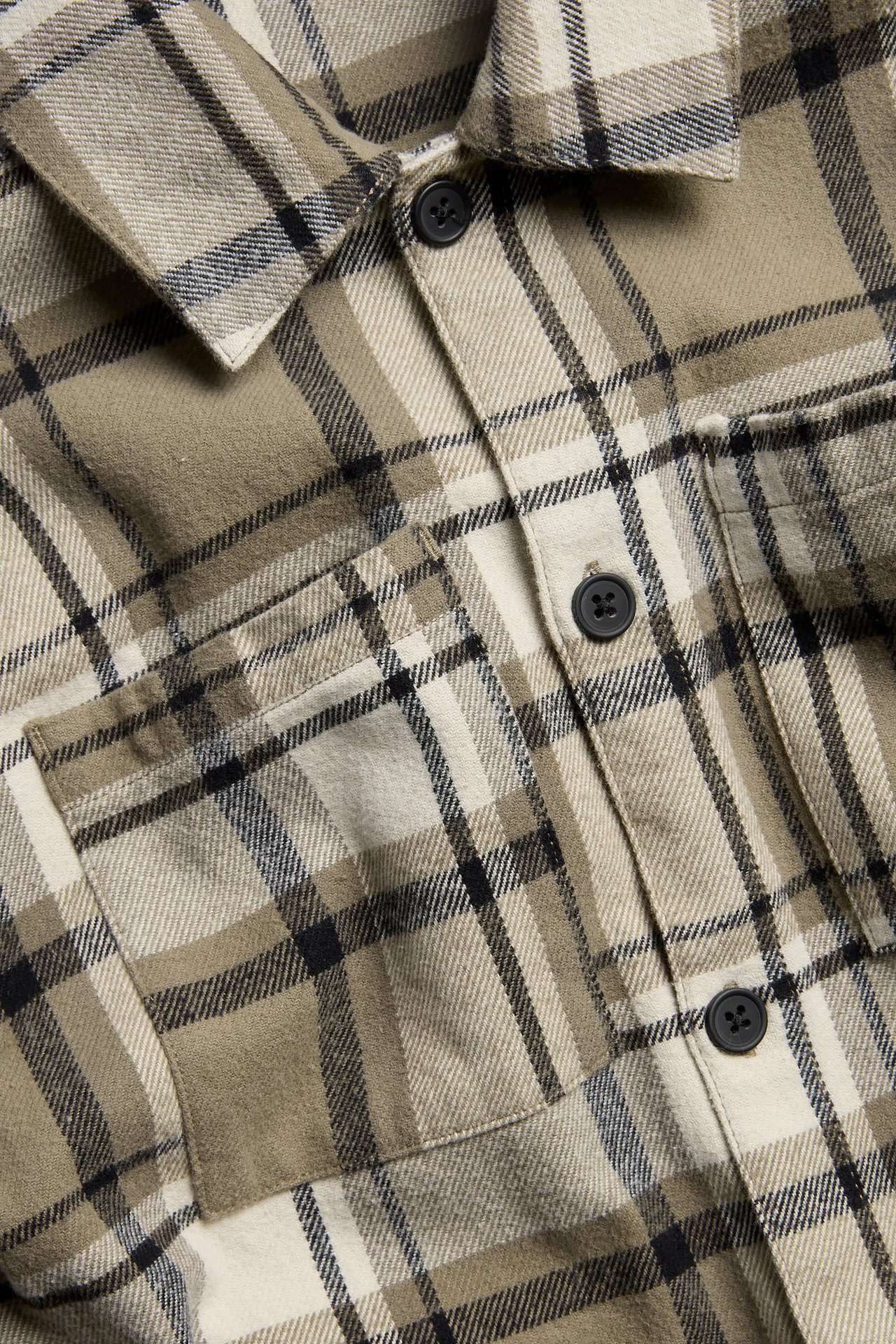 Zbliżenie na beżową koszulę w kratę z flaneli dla dzieci, z czarnymi guzikami i kieszeniami na piersi.