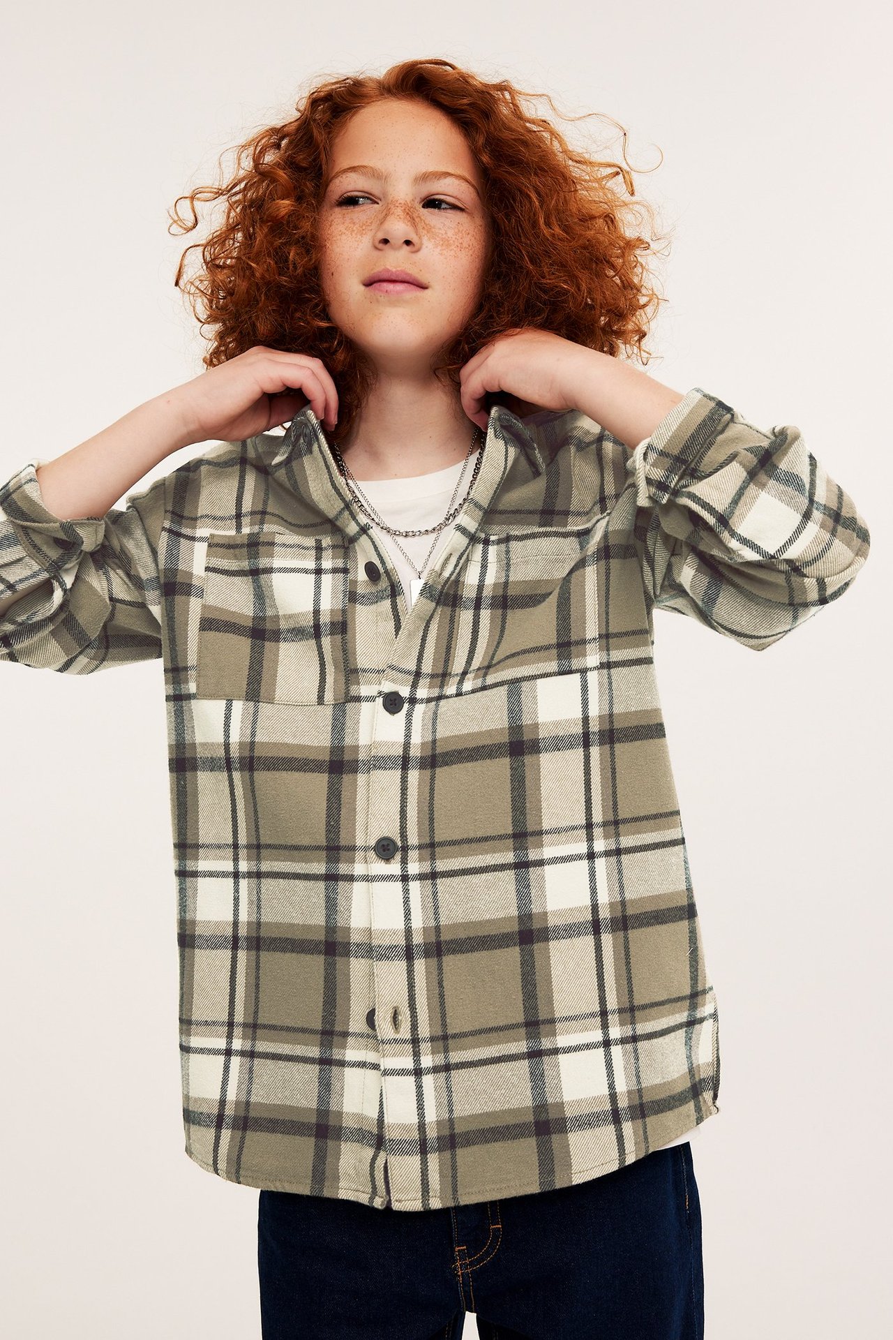 Framifrån: Barn i mörkbeige rutig flanellöverskjorta över vit t-shirt, silverhalsband.