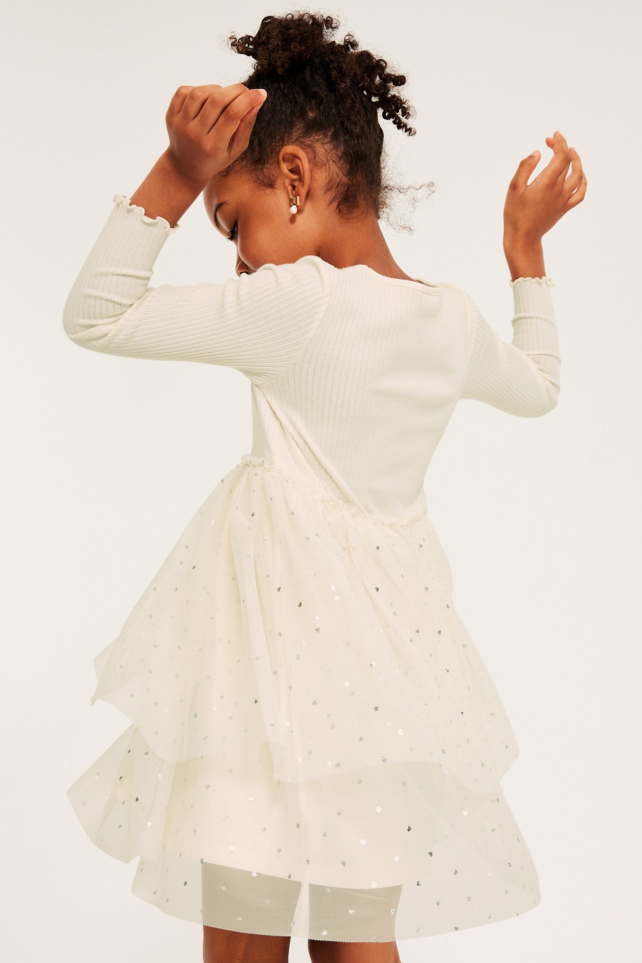 Ryggvy: barn i benvit klänning med ribbad topp, skiktad tyllkjol mönstrad med silverhjärtan.