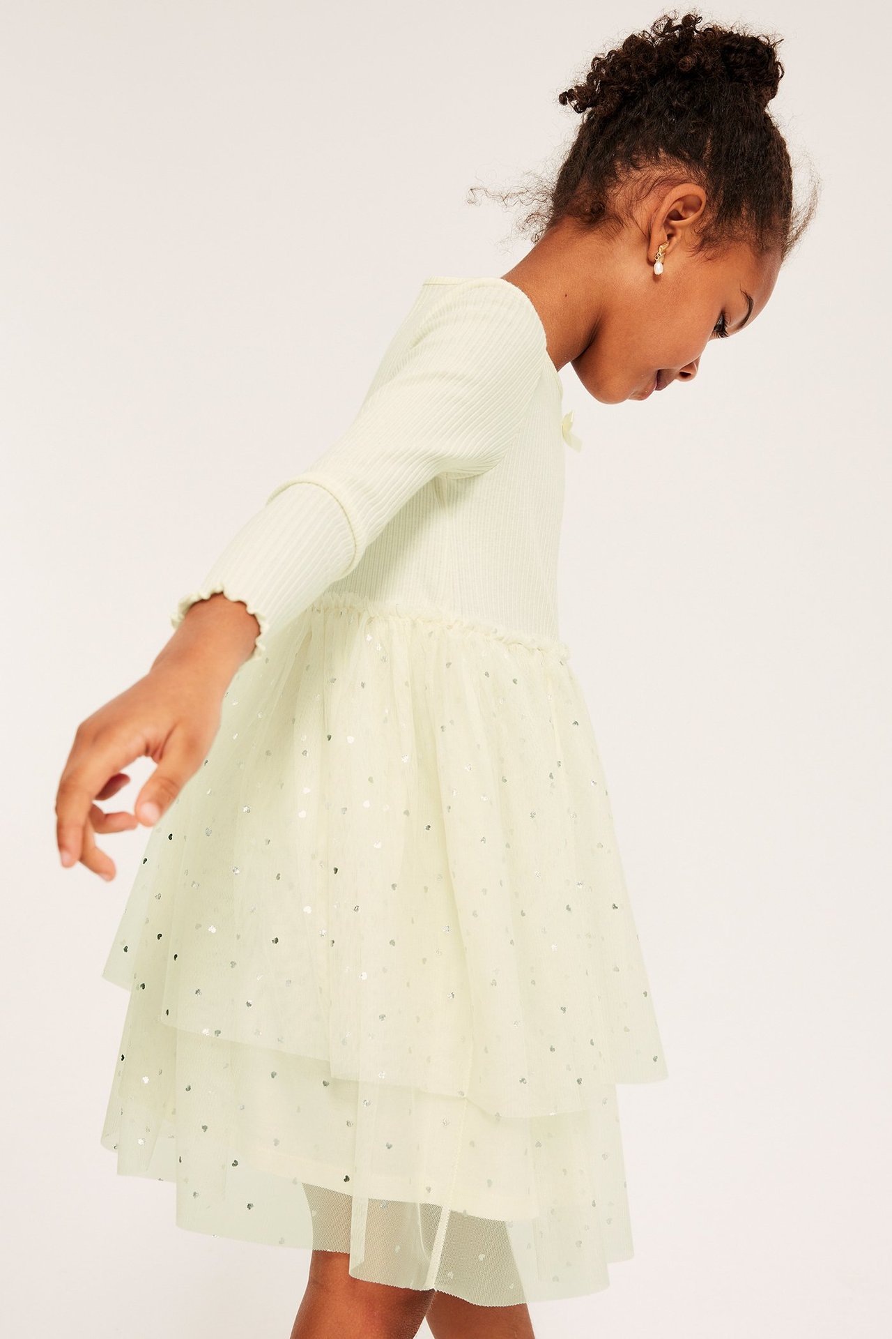 Barnklänning i offwhite med trikå, silverprintad tyllkjol, mönstrad, kort ärm, modell i profil.