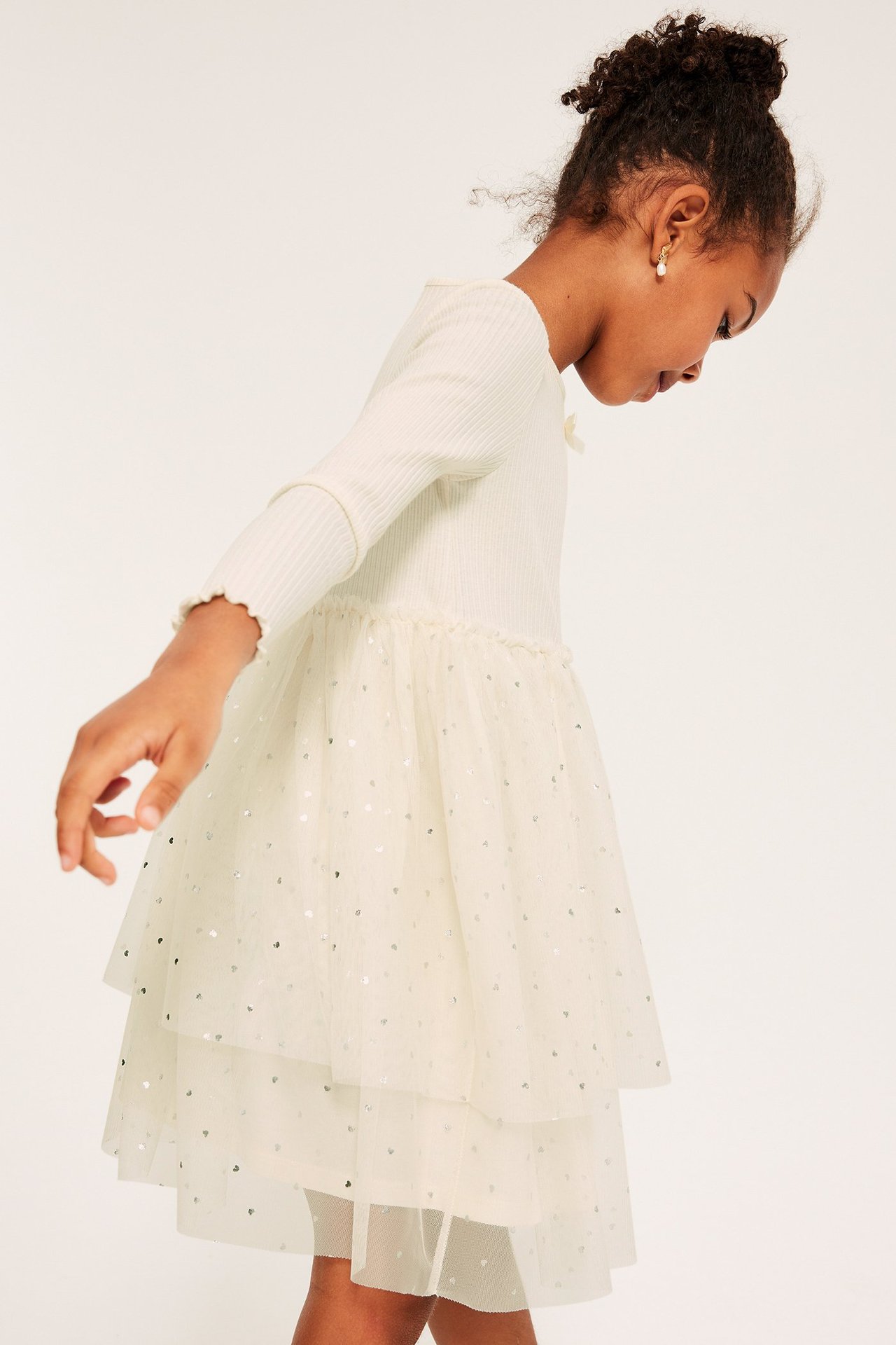 Sideutsikt av barn i offwhite ribbet kjole med glitrende tyllskjørt i flere lag og perleøredobb.