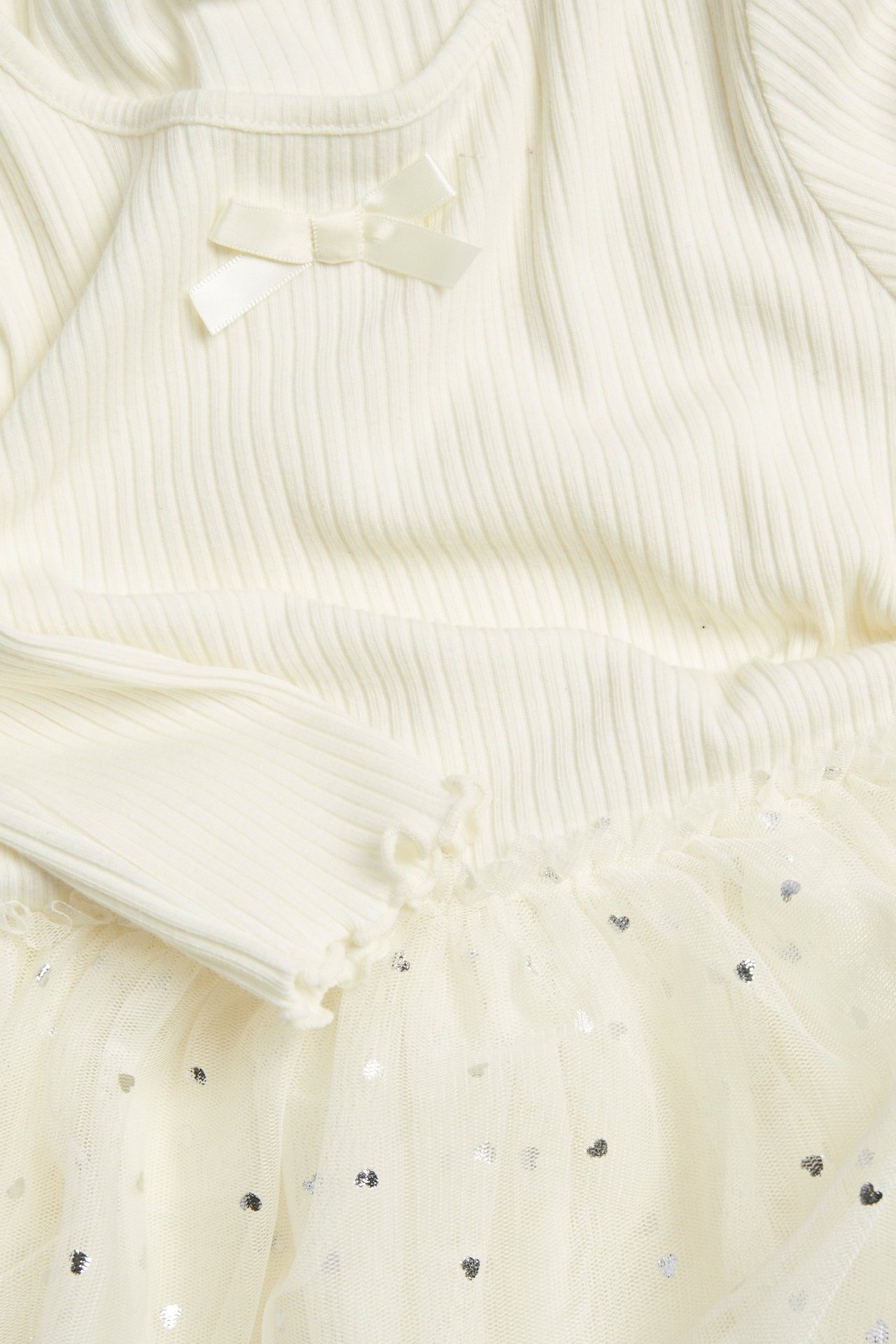 Zbliżenie na ecru, prążkowaną sukienkę dziecięcą z kokardką i tiulową spódnicą w srebrne serduszka.