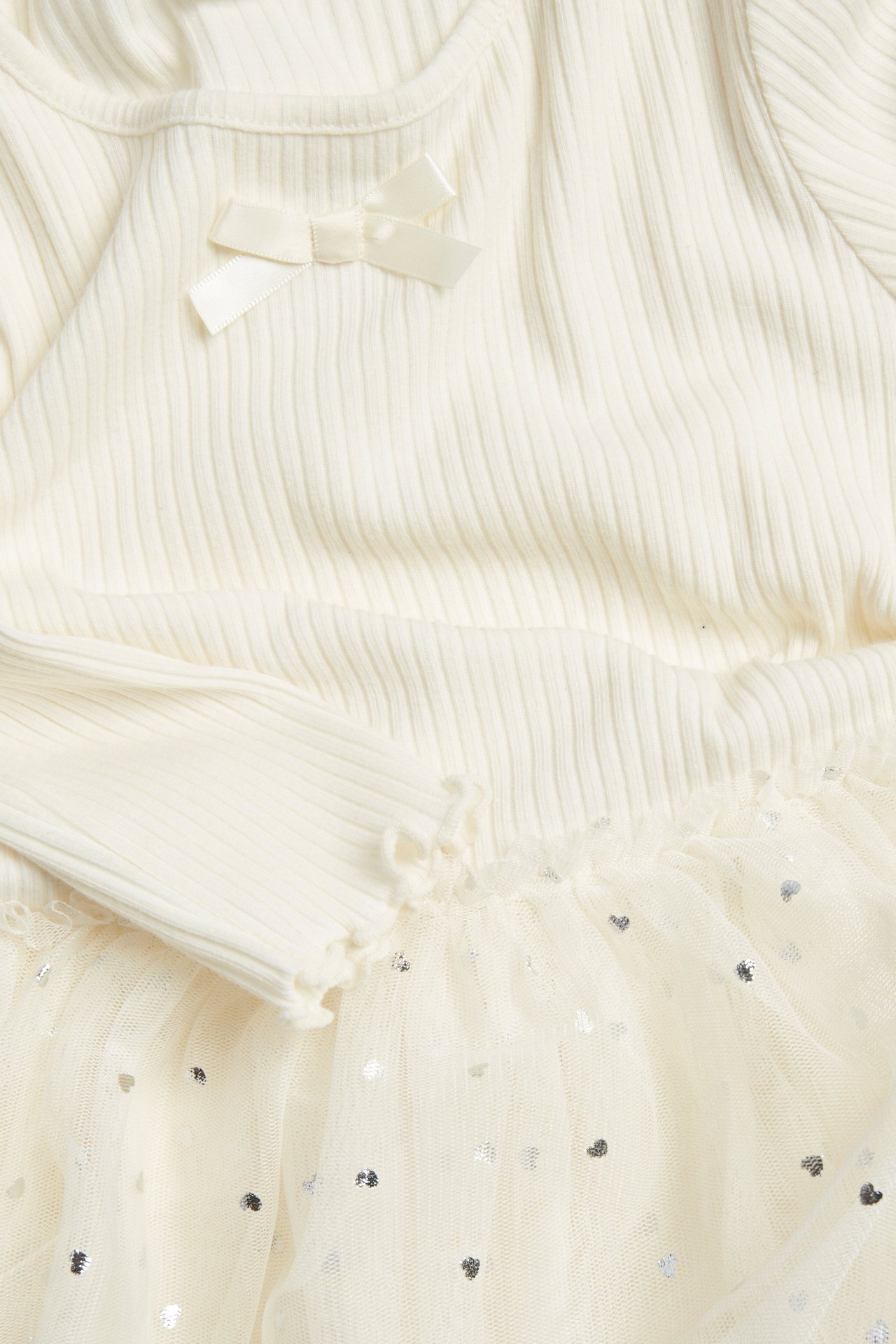 Zbliżenie: Dziecięca sukienka off-white z prążkowaną górą, kokardką, długimi rękawami i tiulową spódnicą w serca.