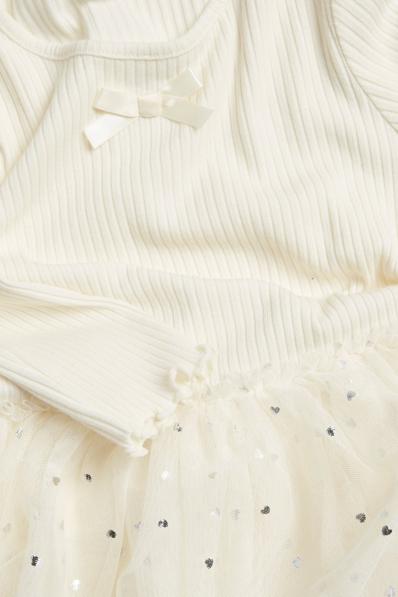 Zbliżenie: Dziecięca sukienka off-white z prążkowaną górą, kokardką, długimi rękawami i tiulową spódnicą w serca.