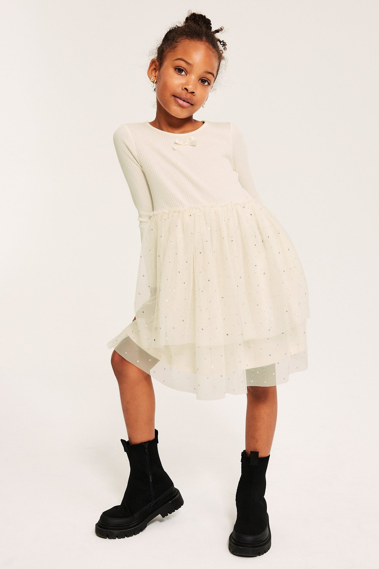Widok z przodu dziecka w kremowej prążkowanej sukience z tiulową spódnicą w brokatowy wzór i czarnych butach.
