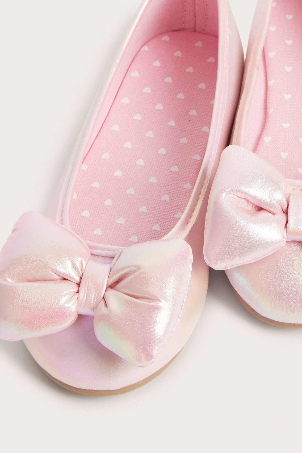Zbliżenie na opalizujące jasnoróżowe balerinki dziecięce z dużymi kokardami i wkładkami w serca.