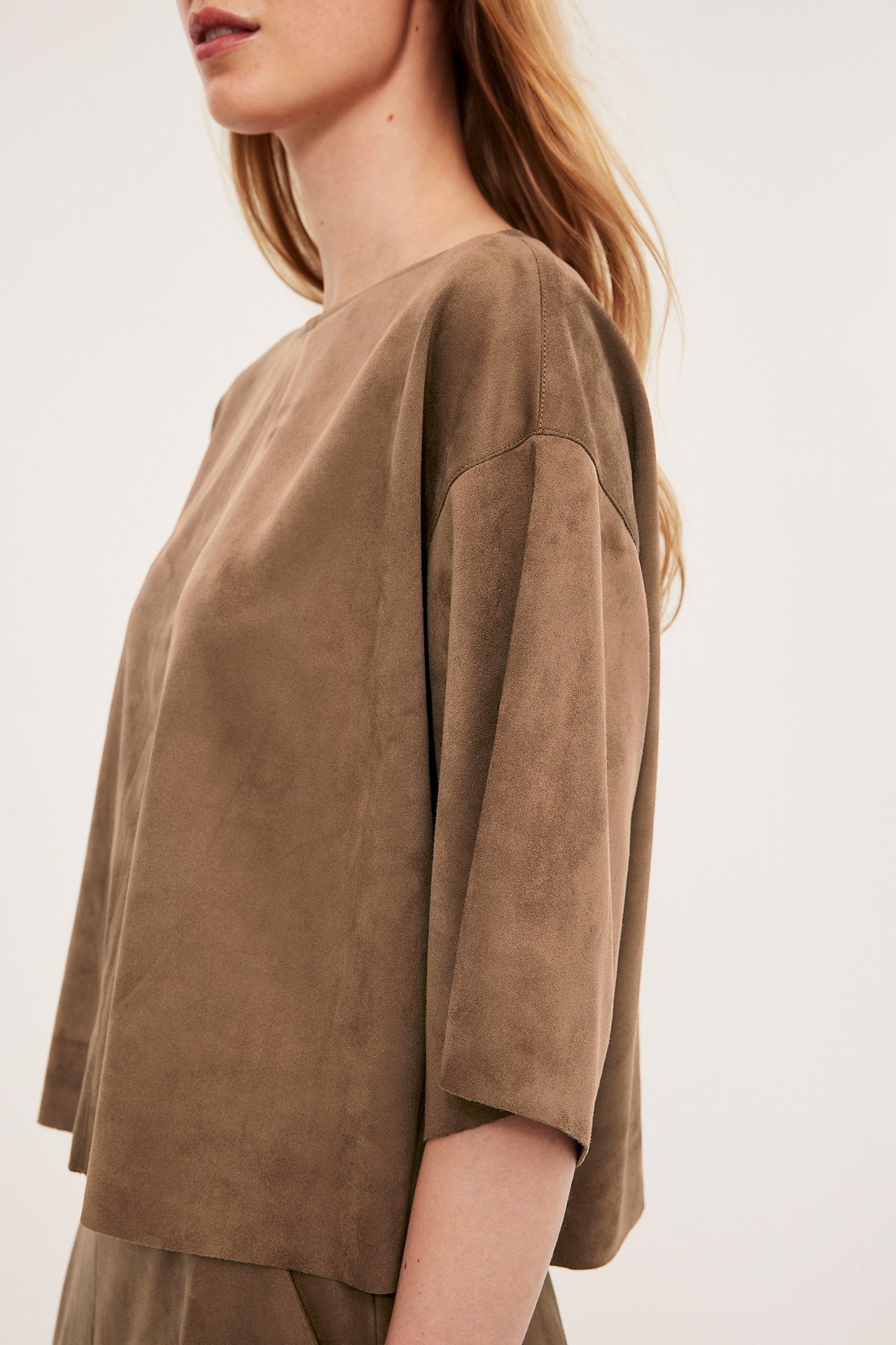 Sidovy av en kvinna i en lös brun blus i mockaimitation med trekvartsärm.