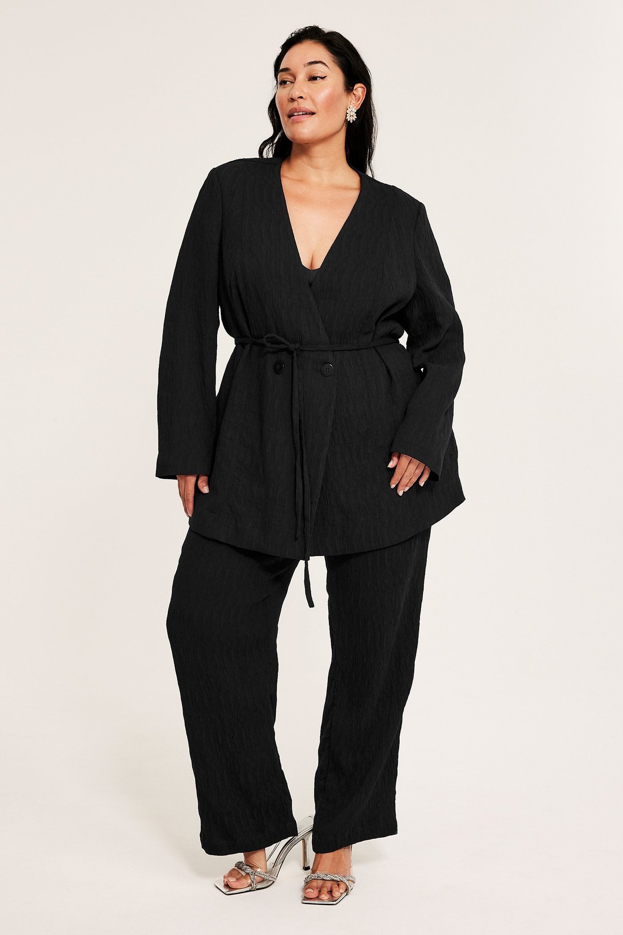 Widok z przodu: Kobieta w czarnej teksturowanej marynarce z wiązaniem, pasujących spodniach i srebrnych sandałach.