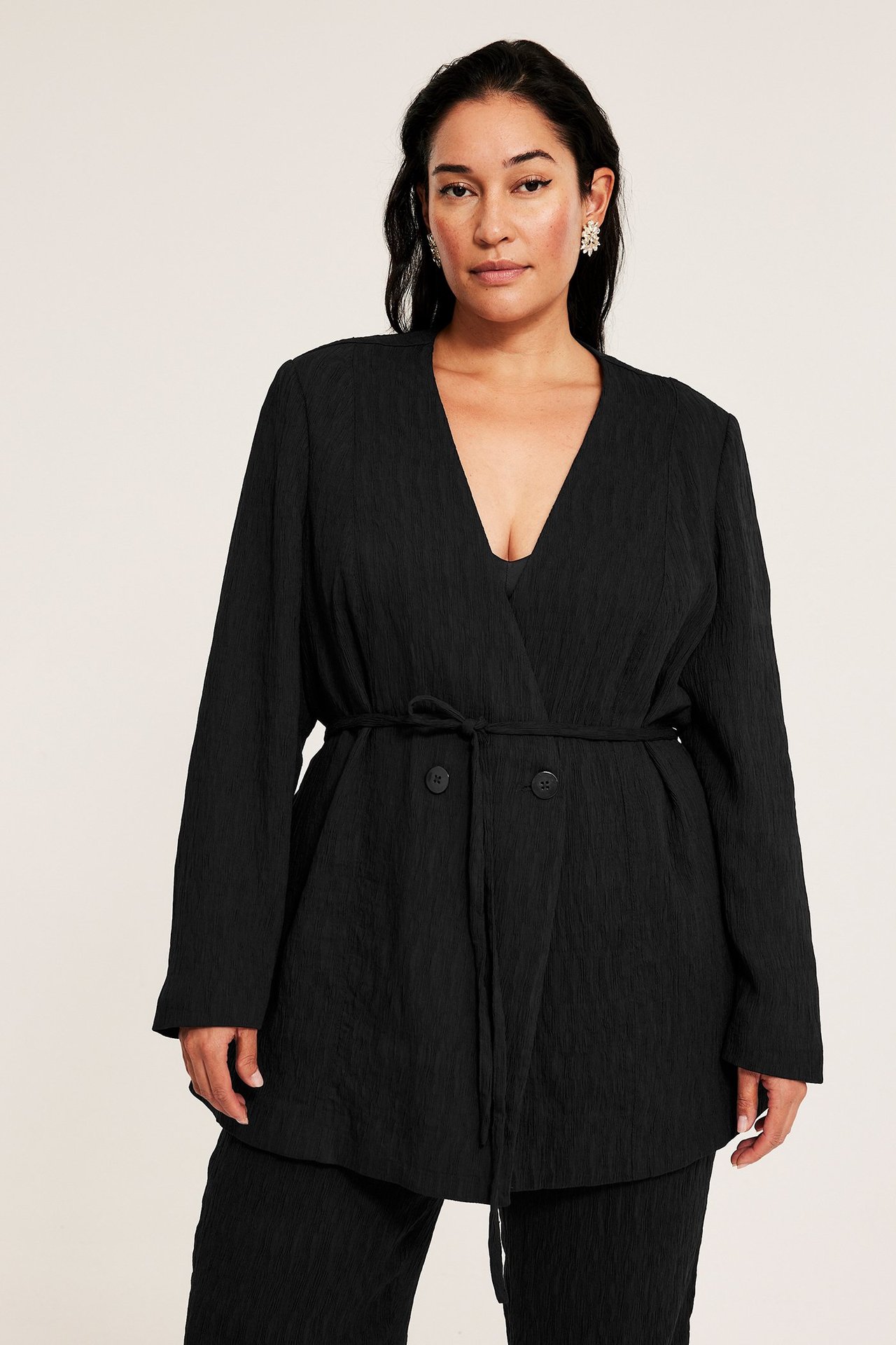 Widok z przodu: Kobieta w czarnej teksturowanej marynarce z wiązaniem, na czarnym topie i spodniach.