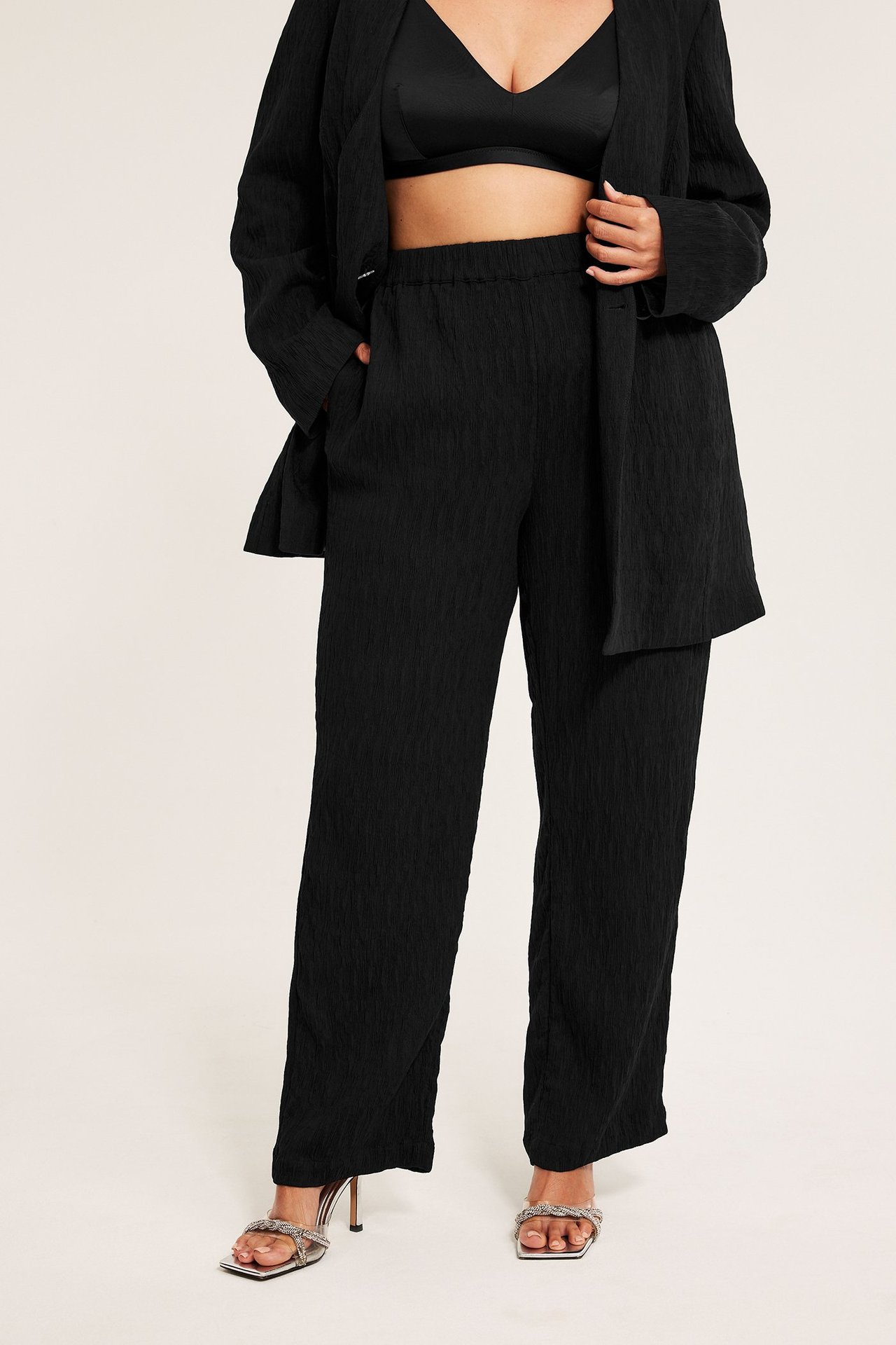 Framifrån: kvinna i svarta texturerade pull on-byxor, matchande kavaj, svart bralette och silversandaler.