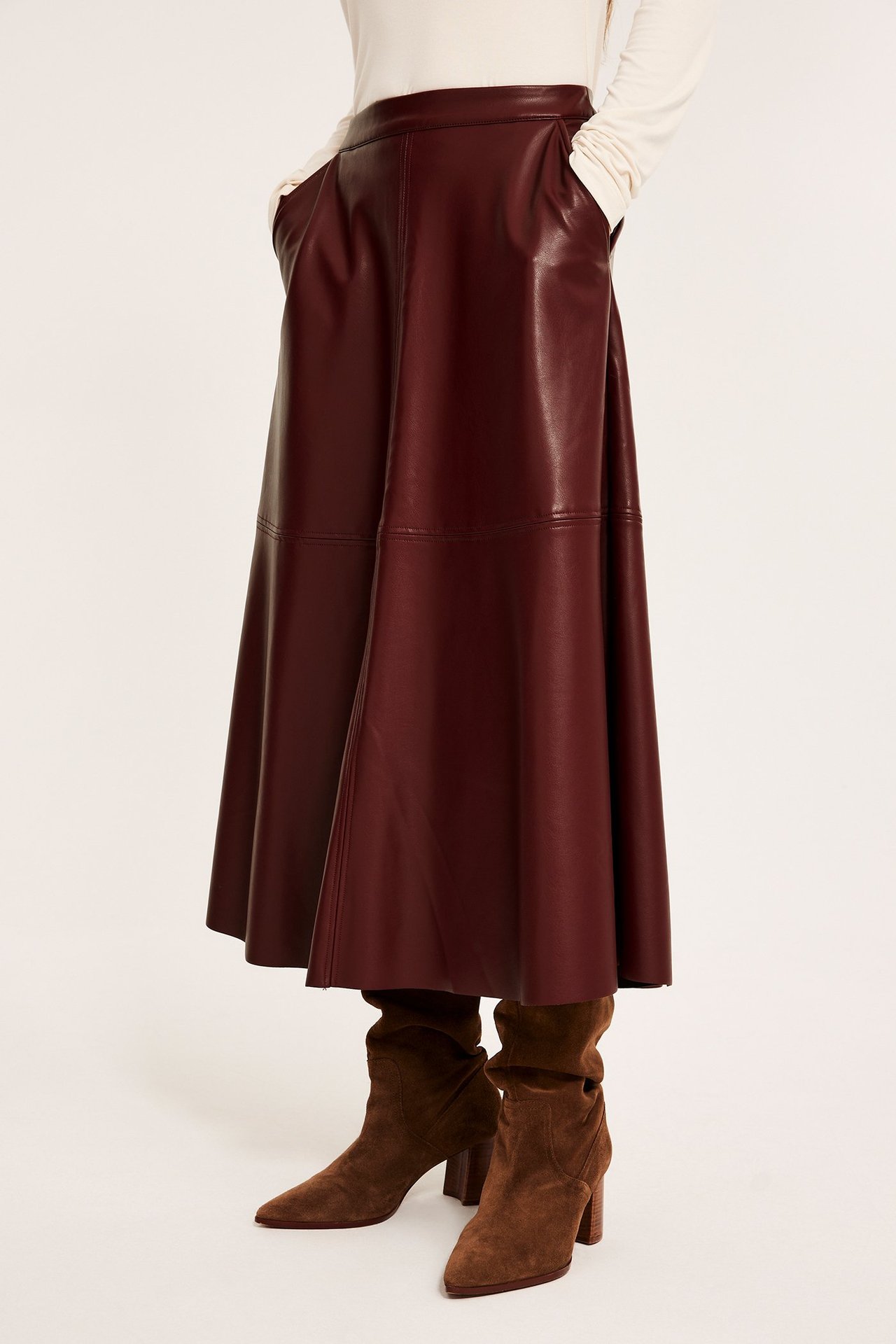 Naisen edestä: pitkä, levenevä punainen tekonahkahame taskuilla, yhdistettynä kermanväriseen pitkähihaiseen toppiin ja ruskeisiin mokkanahkasaappaisiin.