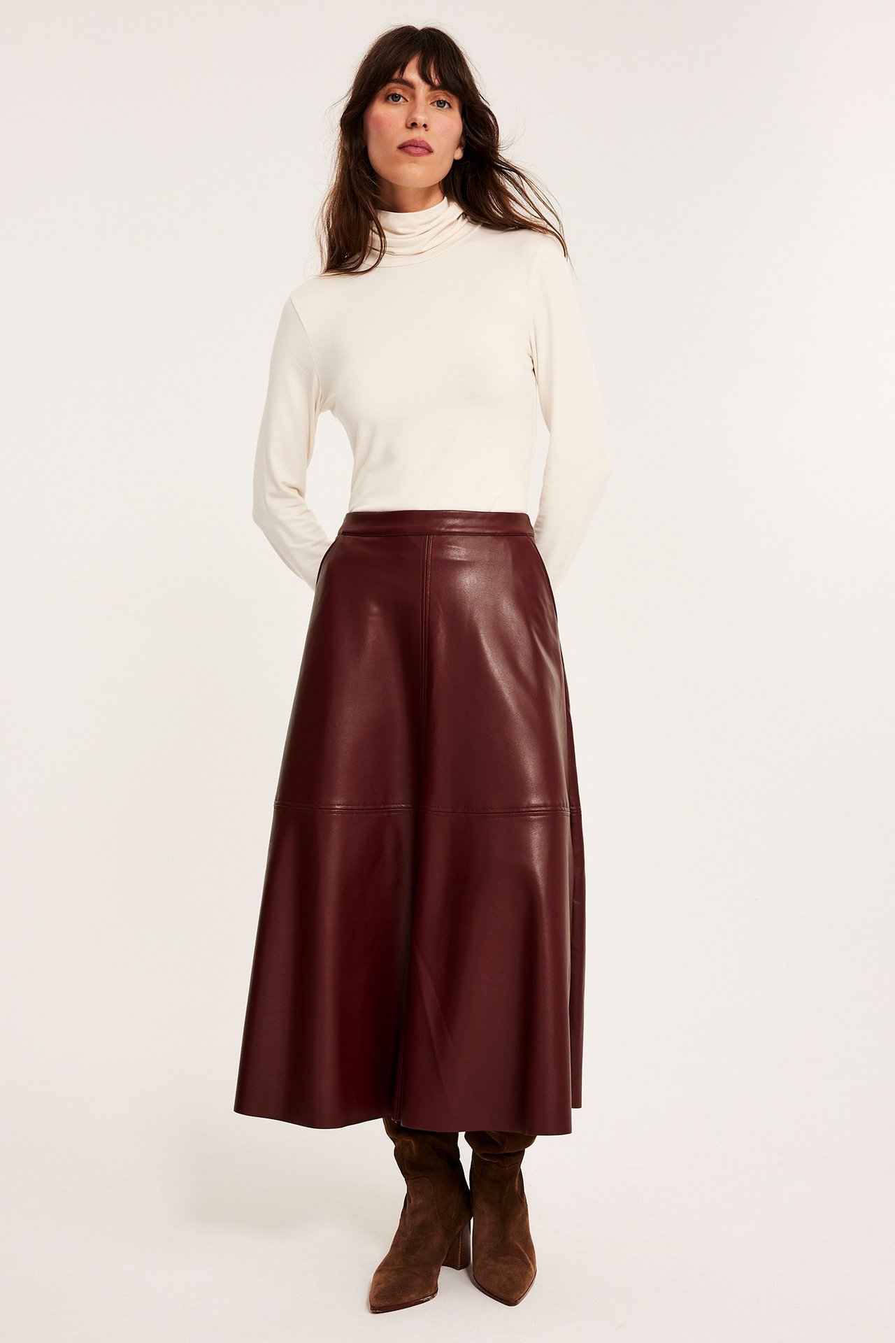 Edestä kuvattu nainen pitkässä, levenevässä punaisessa tekonahkahameessa, kermanvärisessä poolopaidassa ja ruskeissa mokkanahkasaappaissa.