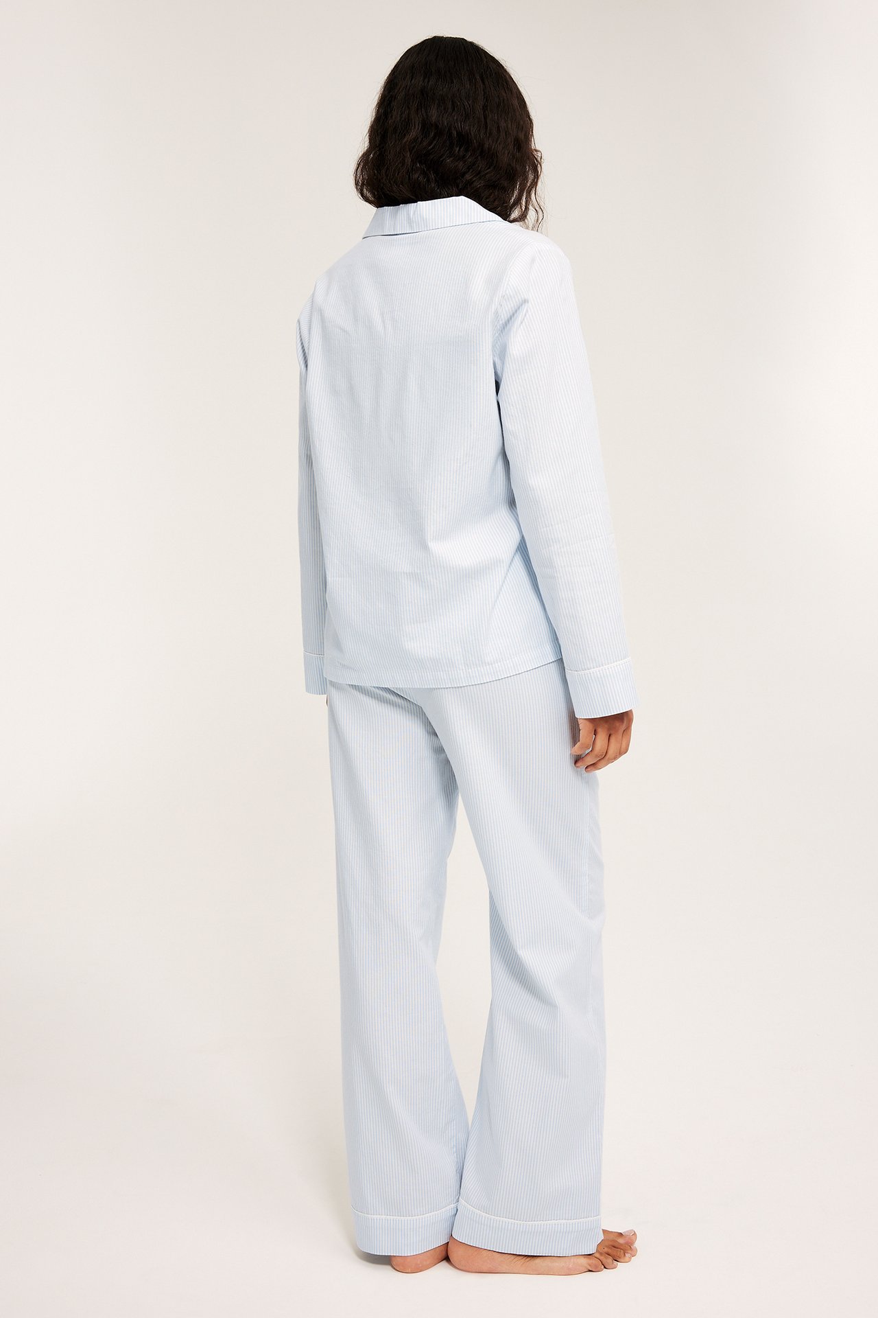 Bakfra: Kvinne i lyseblå stripete bomullspysjamas med lange ermer og bukser med hvit piping.