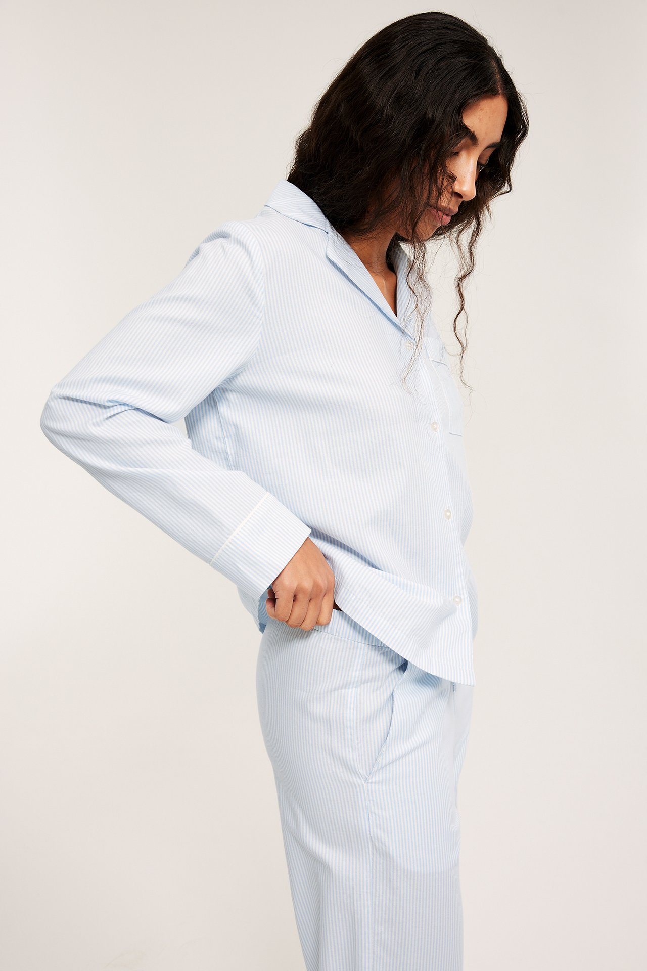 Sidebilde av en kvinne i et lyseblått stripete vevd pyjamassett med passpoaldetaljer.