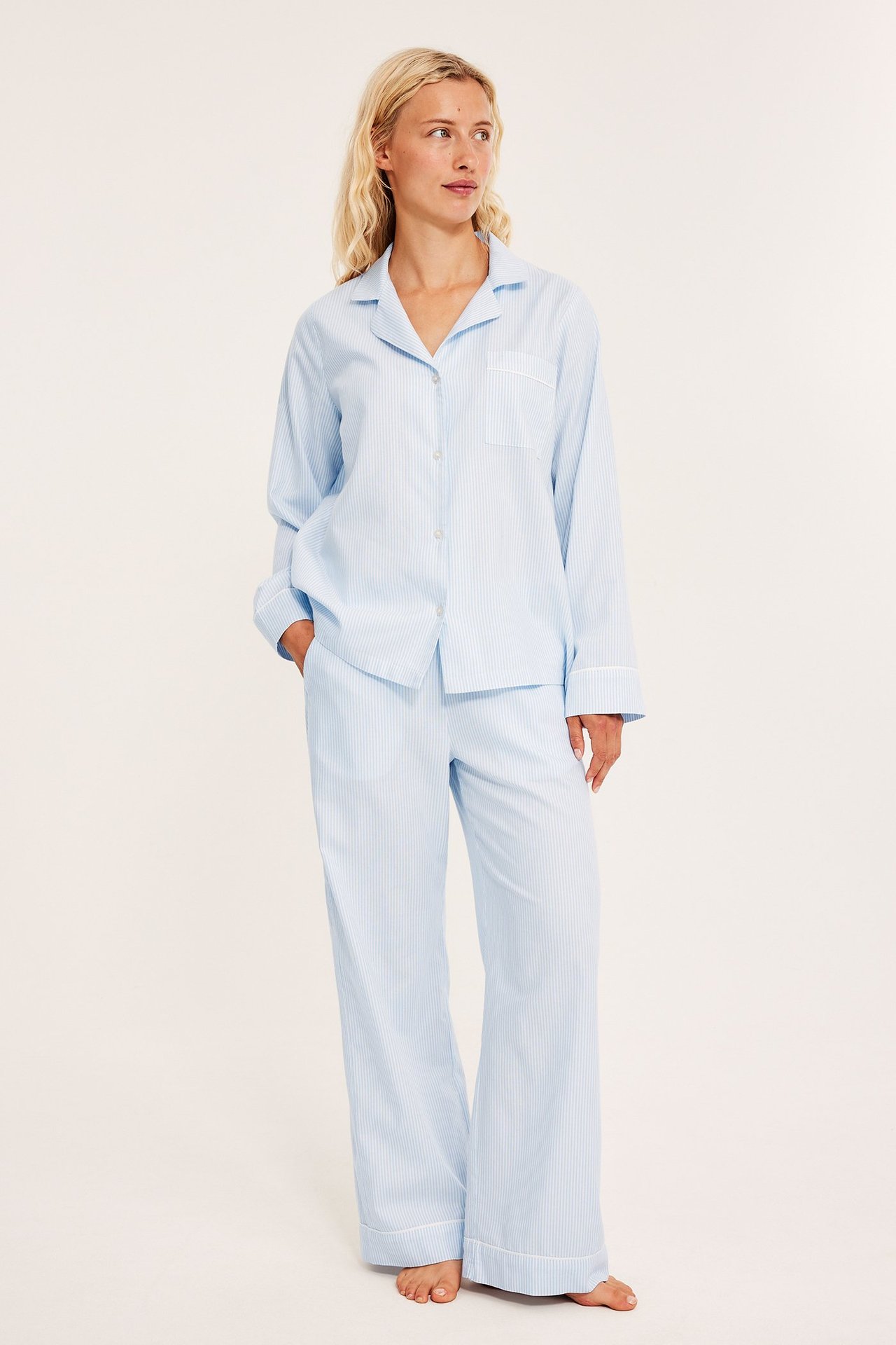 Edestä näkymä: Nainen yllään vaaleansininen raidallinen puuvillapyjama valkoisella tereellä, kädet taskuissa.