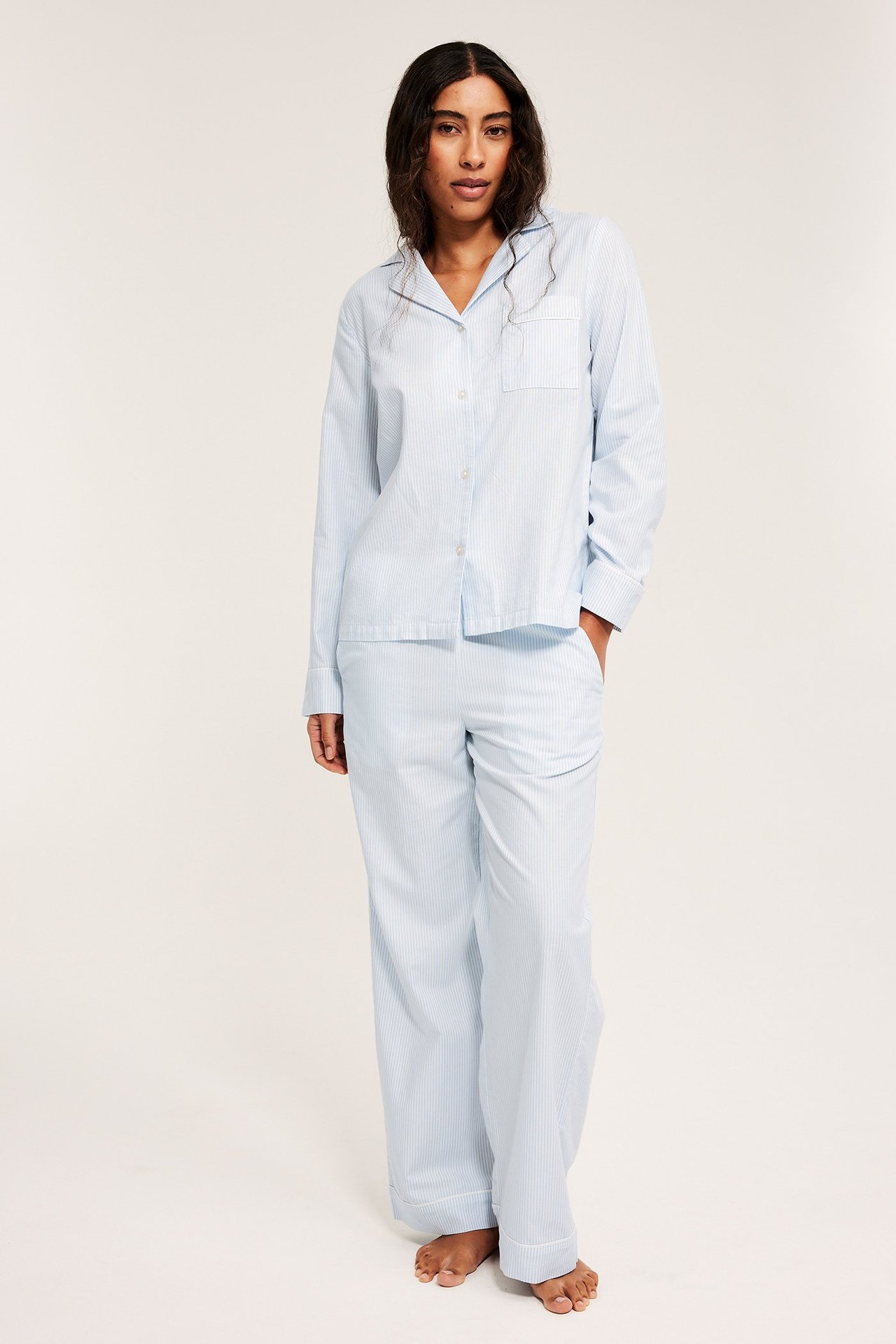 Framsida av en kvinna i ett ljusblårandigt vävt pyjamasset i bomull med passpoaldetaljer.