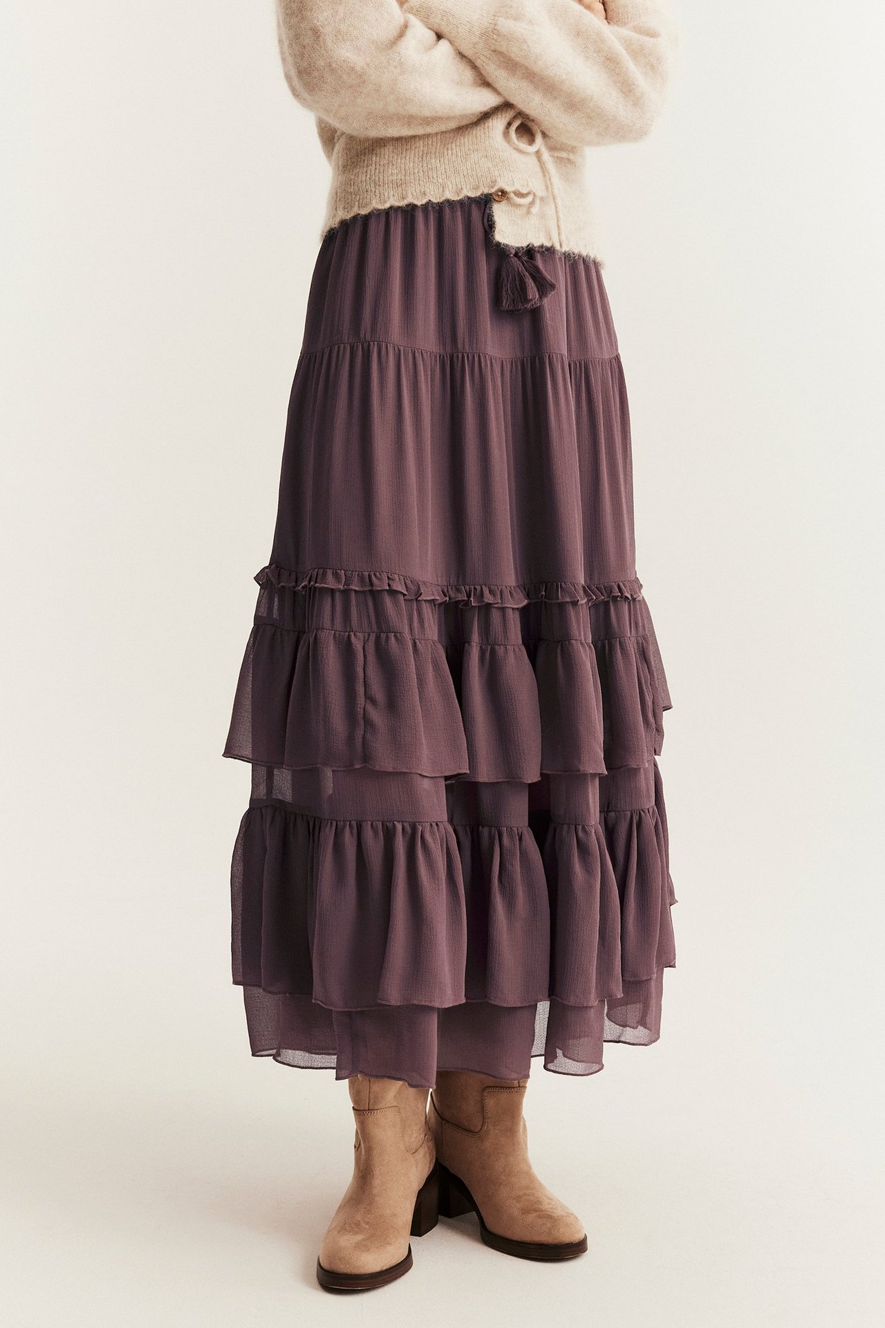 Framifrån, kvinna i lång lila volangkjol i chiffong med tofsar, beige kofta och beige ankelstövlar.