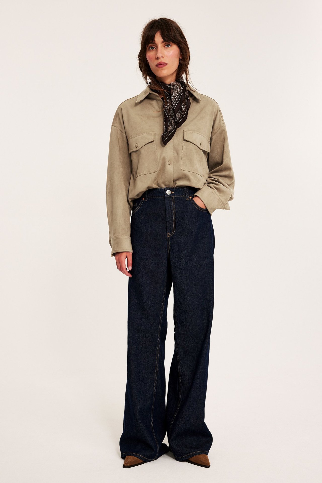 Frontvy: Kvinna i beige skjorta i mockaimitation med bröstfickor, mörkblå jeans, svart bandana.