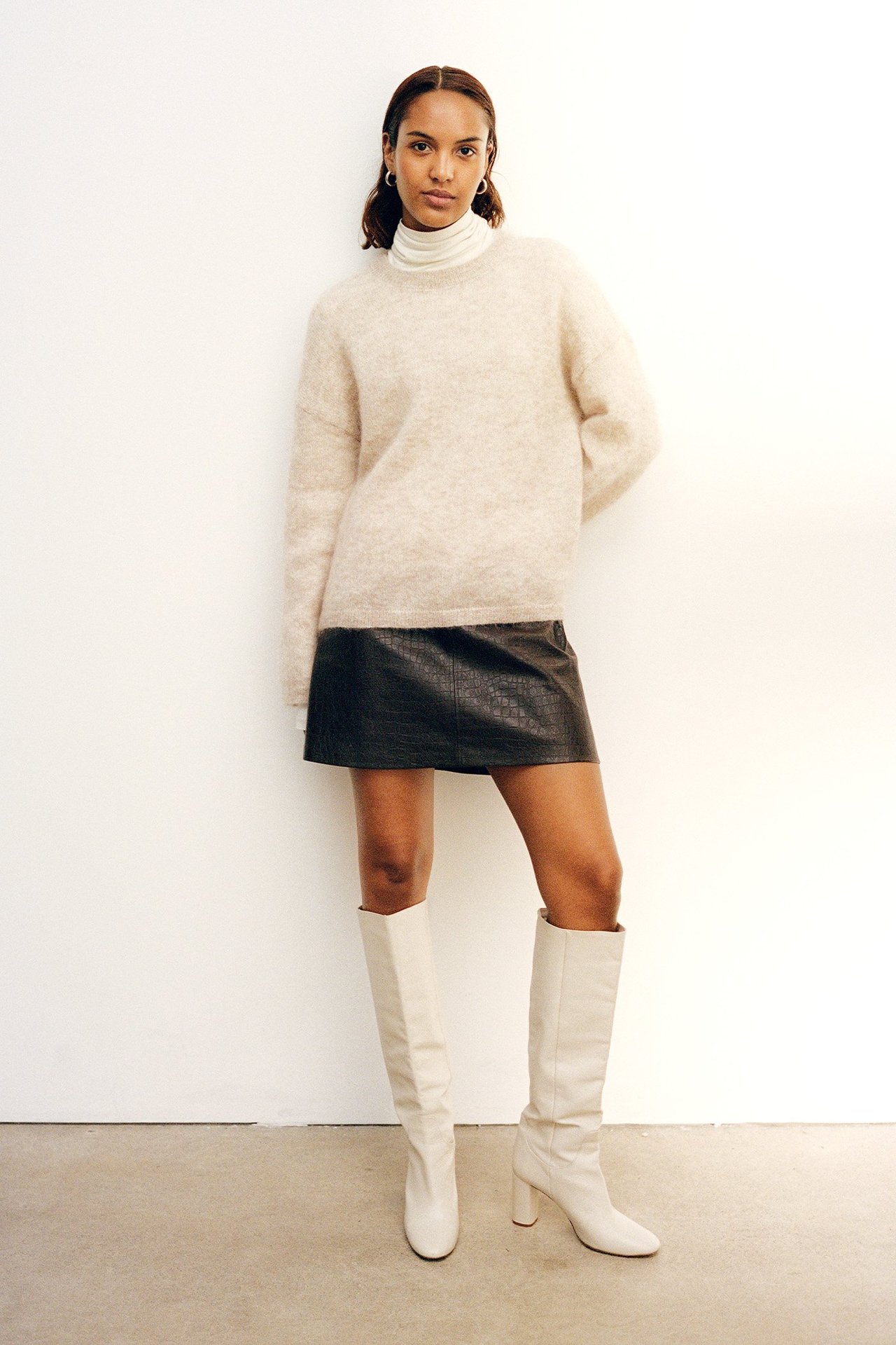 Edestäpäin: nainen yllään tummanruskea lyhyt kuvioitu tekonahkahame, beige neule ja valkoiset saappaat.