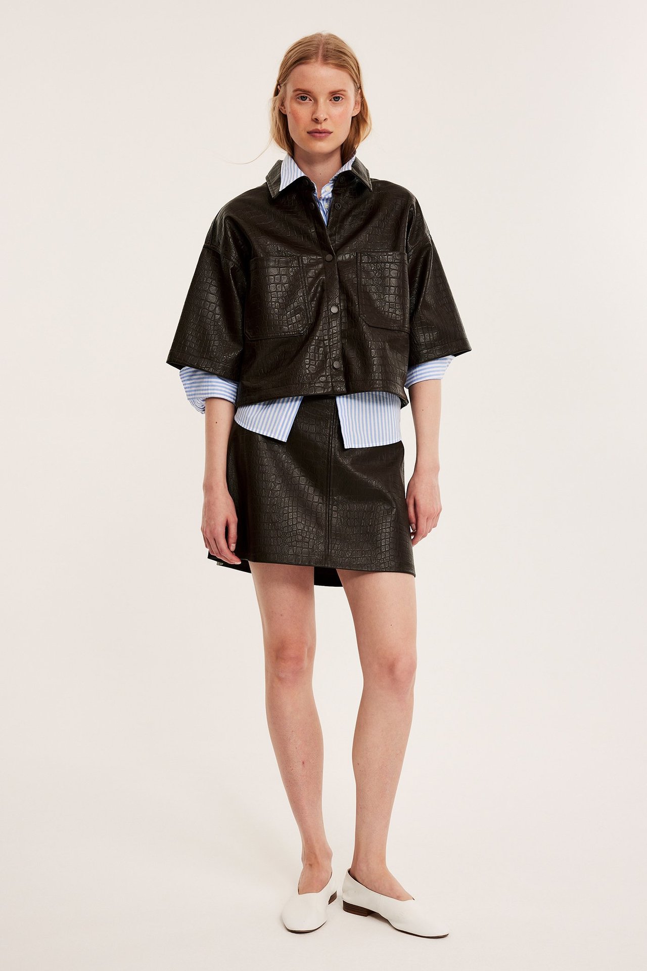 Etunäkymä. Nainen tummanruskeassa krokotiilikuviollisessa tekonahkahameessa, paidassa ja valkoisissa kengissä.