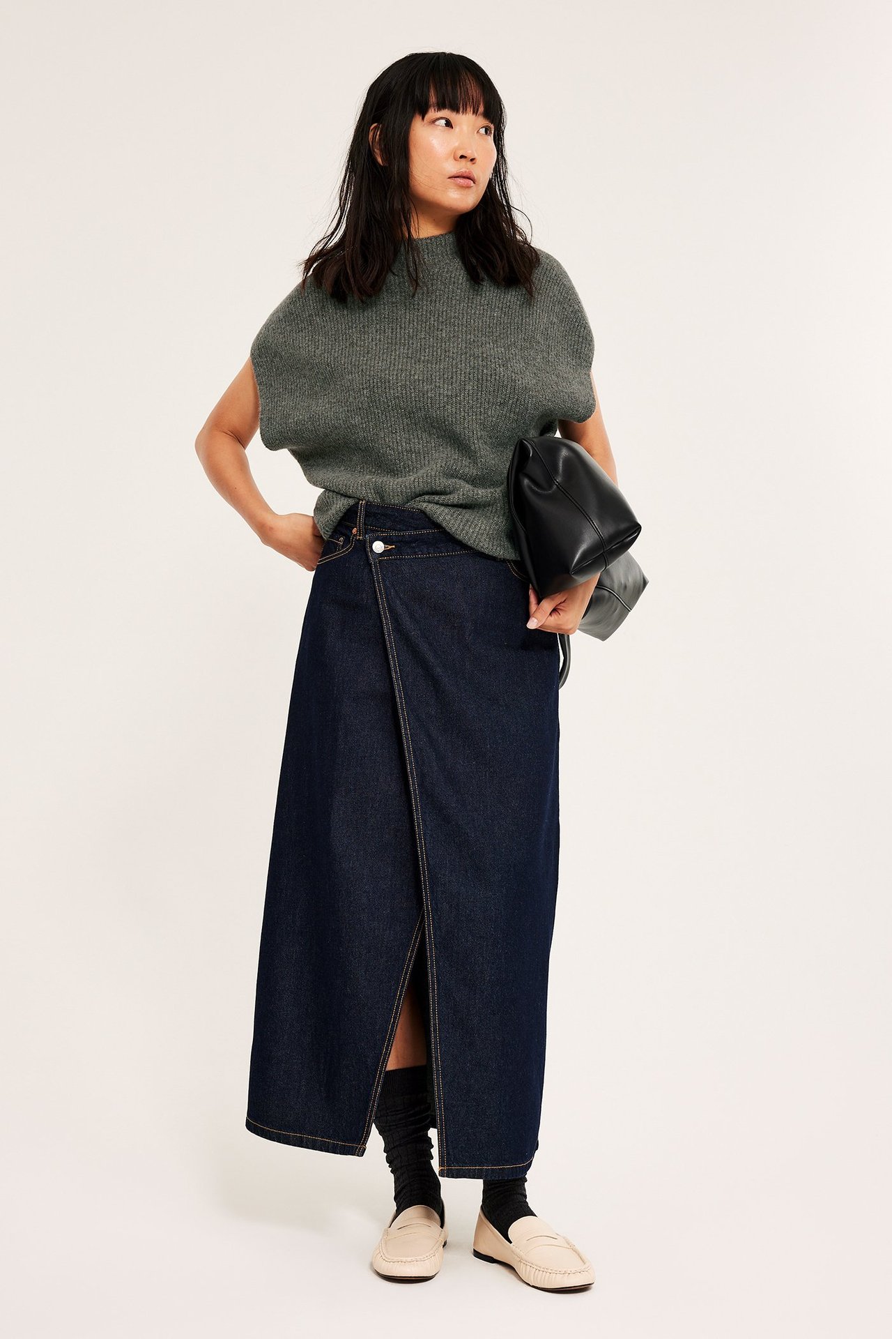 Kvinna bär en ribbstickad väst i brun melange, över tröja, tillsammans med jeanskjol och loafers.