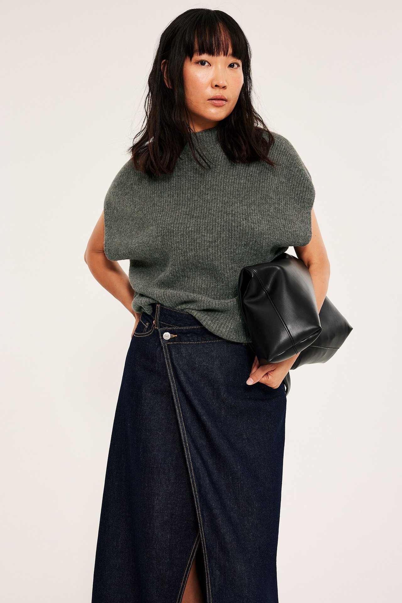 Kvinna i grå ribbstickad väst, svart kjol, håller svart handbagage, frontvy.