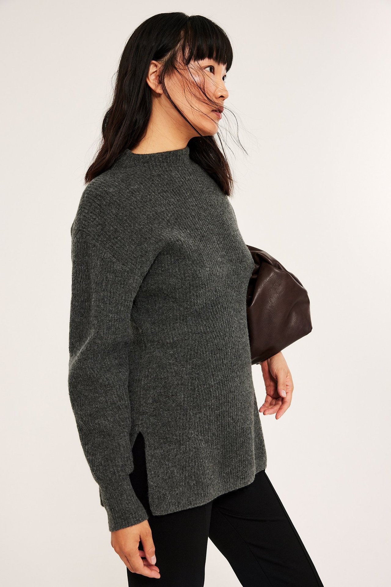 Naisen sivukuva tummanharmaassa ribbineulepaidassa puolipoolokauluksella, mustissa housuissa, pitäen ruskeaa clutch-laukkua.