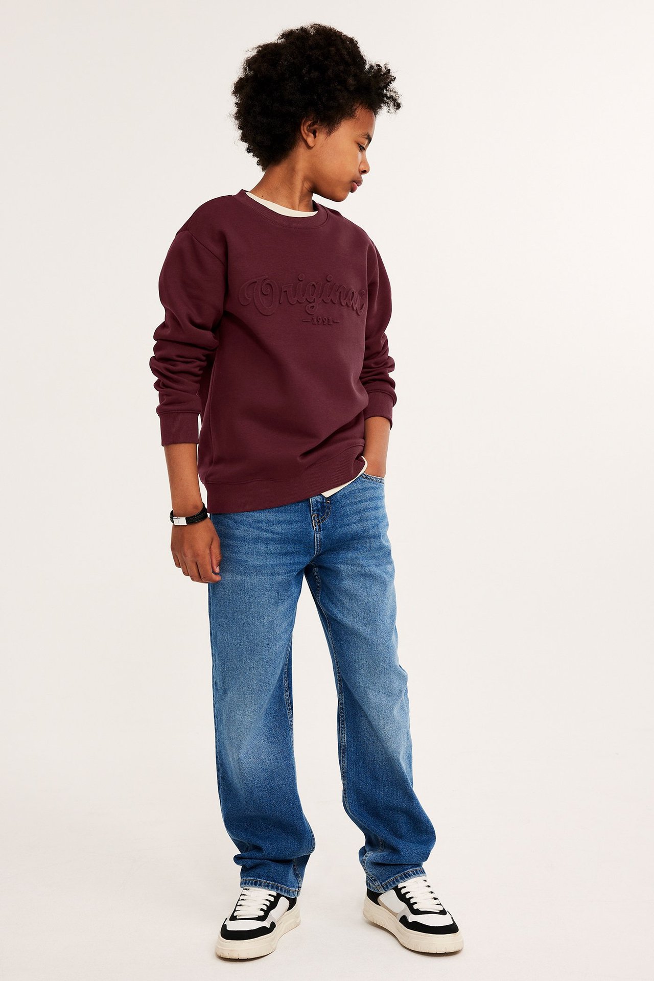 Dziecko z przodu: ciemnoczerwona bluza z nadrukiem 'Original', niebieskie dżinsy i białe trampki.