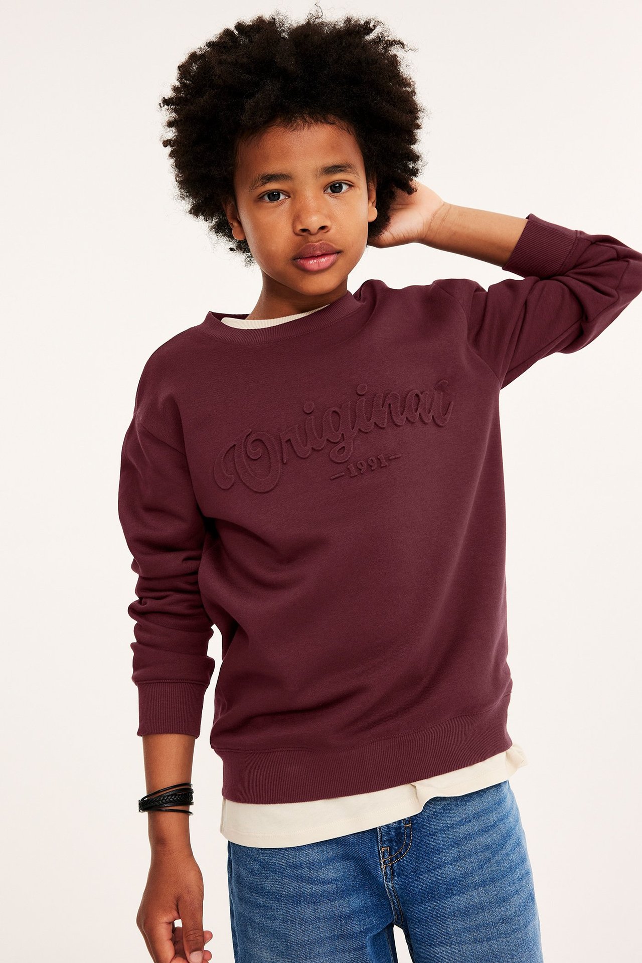 Widok z przodu dziecka w ciemnoczerwonej bluzie z nadrukiem 'Original', niebieskich dżinsach i czarnej bransoletce.