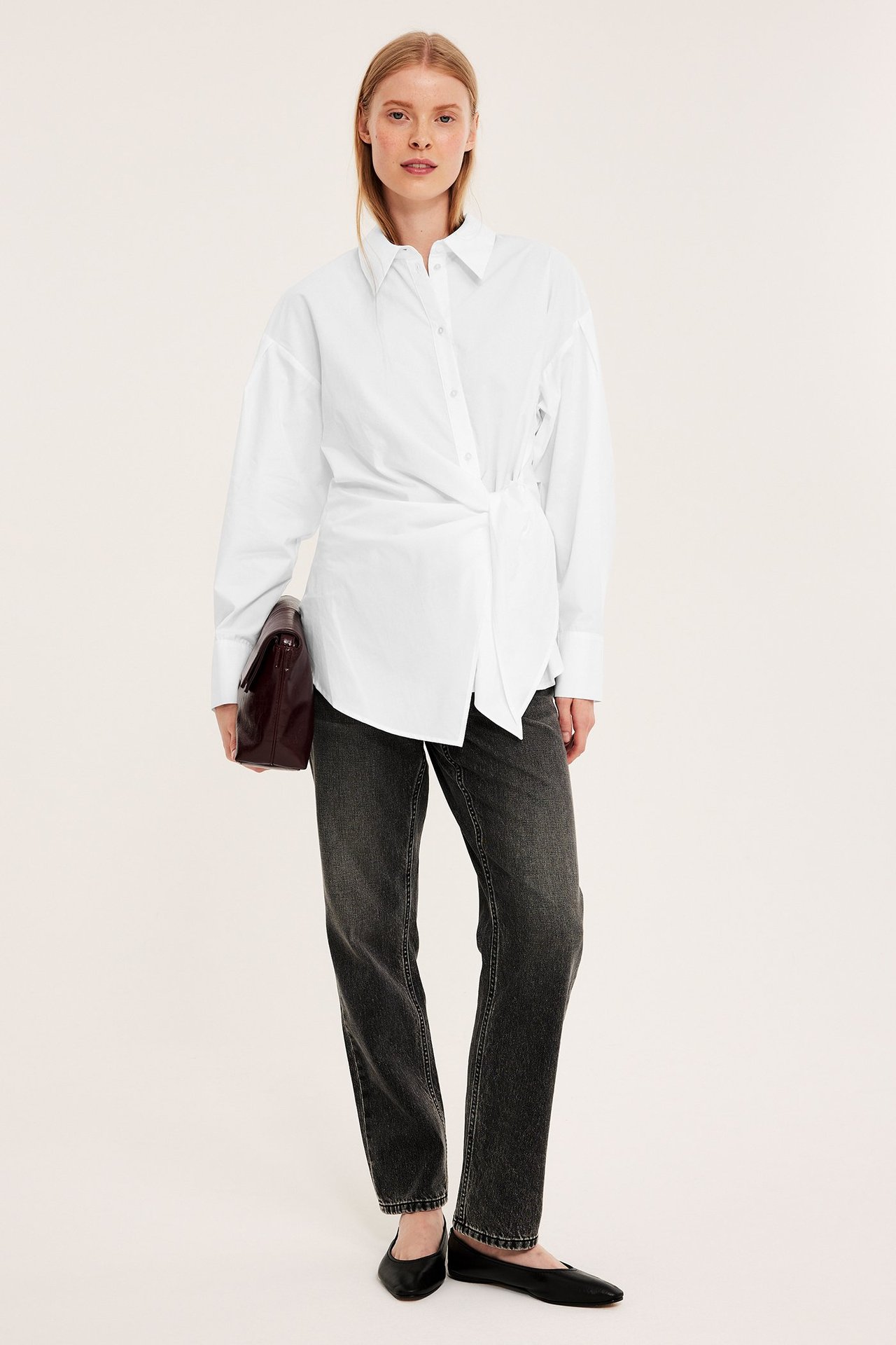Framifrån: Kvinna i vit bomullsskjorta med knytning fram, mörkgrå jeans, svarta ballerinaskor, håller vinröd clutch.